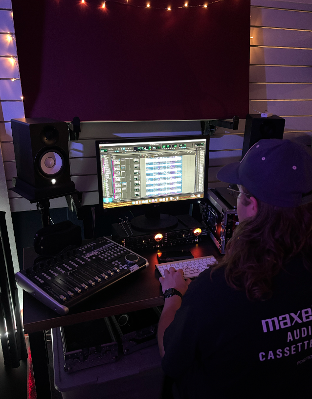 Dallas Lander sitting at a music production workstation with a computer monitor displaying audio editing software, speakers and audio equipment in a studio.