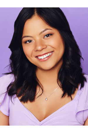 A young woman with long dark hair smiling, wearing a lavender top and a necklace with a heart-shaped pendant, against a purple background.