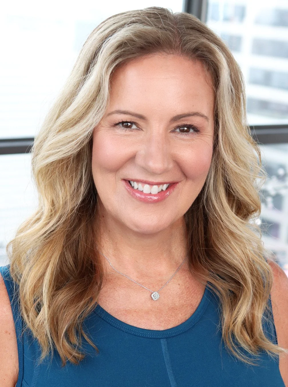 A woman with blonde, wavy hair smiling in front of a window, wearing a blue sleeveless top and a silver necklace.