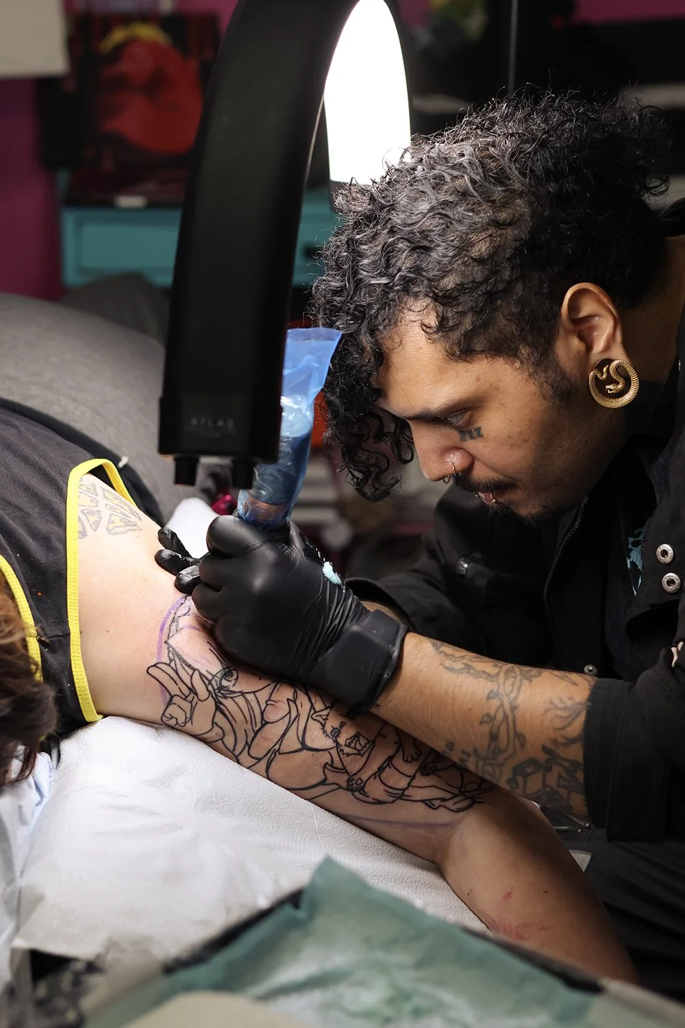 A tattoo artist working on a tattoo on a person's arm with a tattoo machine, wearing black gloves and focused on the work.