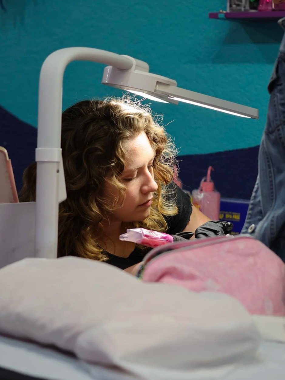 A woman with curly hair receiving a tattoo on her forearm from a tattoo artist in a studio with a blue wall.