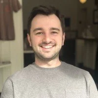 A young man with dark hair and a beard smiling, wearing a gray shirt, standing indoors with a kitchen in the background.