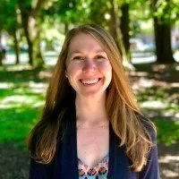 A smiling woman with long hair standing outdoors in a park with trees and sunlight.