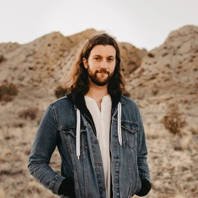 A man with long hair and a beard standing outdoors in a desert landscape, wearing a denim jacket over a white shirt.