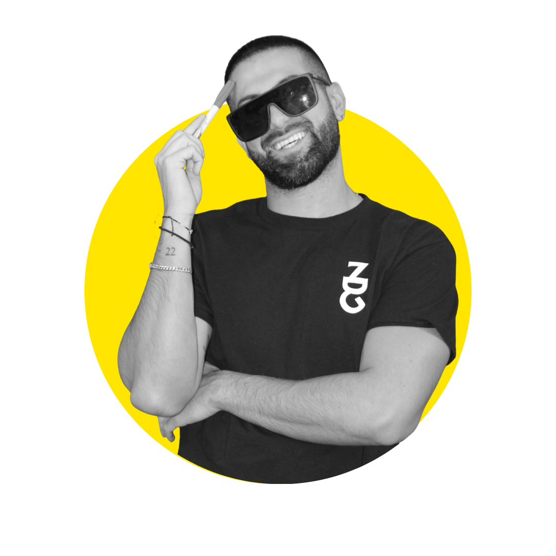 A smiling man with a beard and sunglasses, wearing a black t-shirt with white lettering, is posing with his right hand holding his sunglasses and his left arm crossed.