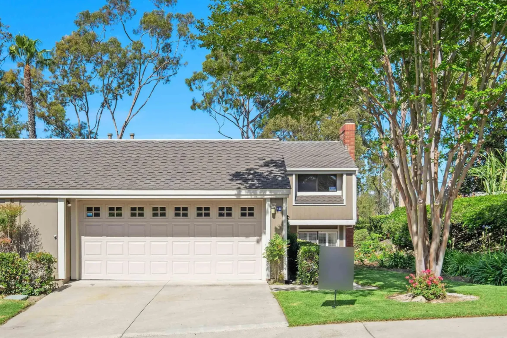 A suburban house with a brown garage door, cream exterior walls, and a gable roof. A large tree with green leaves is on the right, and smaller shrubs and grass are in the front yard. Clear blue sky is above.