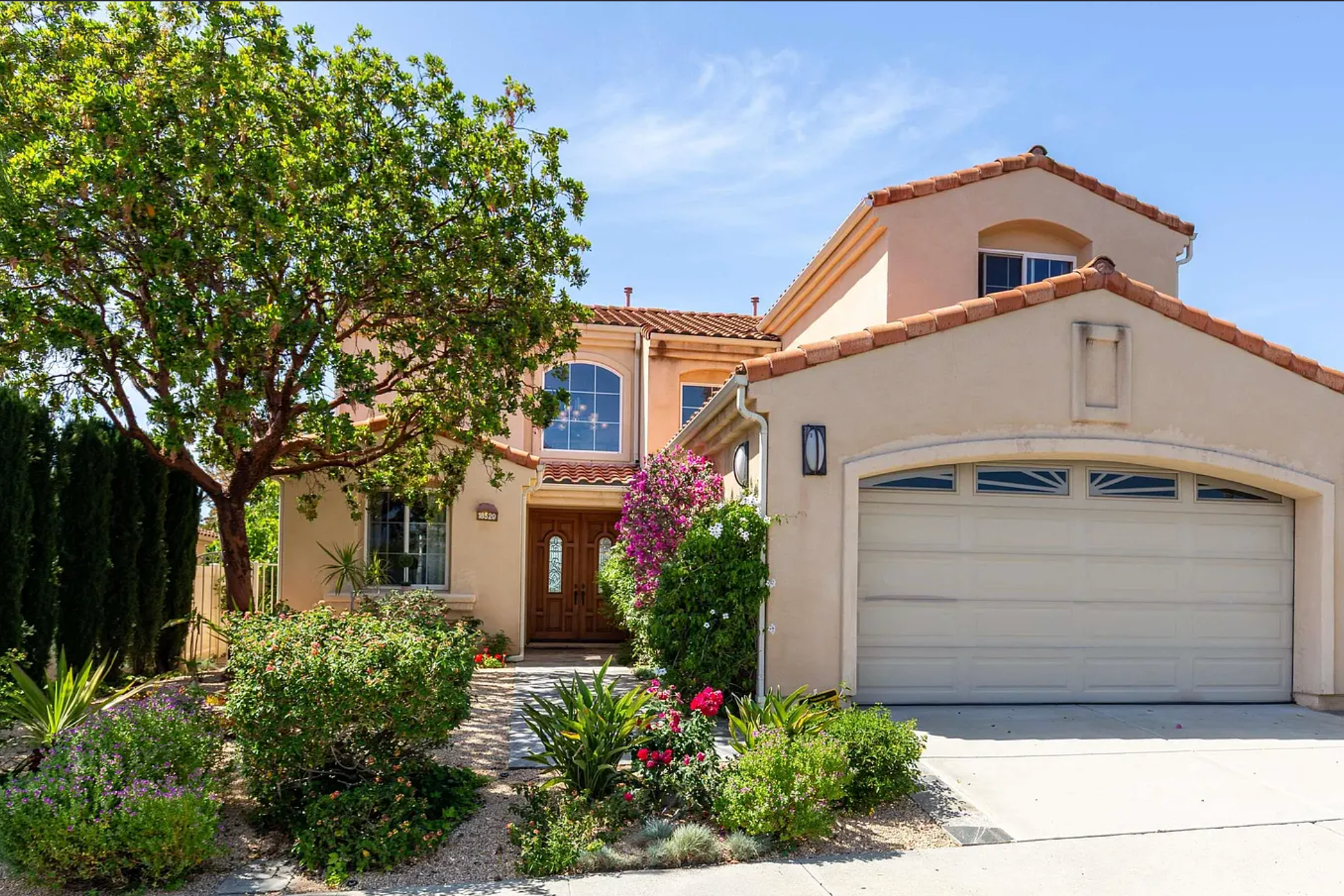 A two-story house with a white garage door, tan stucco walls, and a red tiled roof. There are tall green trees and colorful flowering bushes in the front yard. The sky is clear with some clouds.