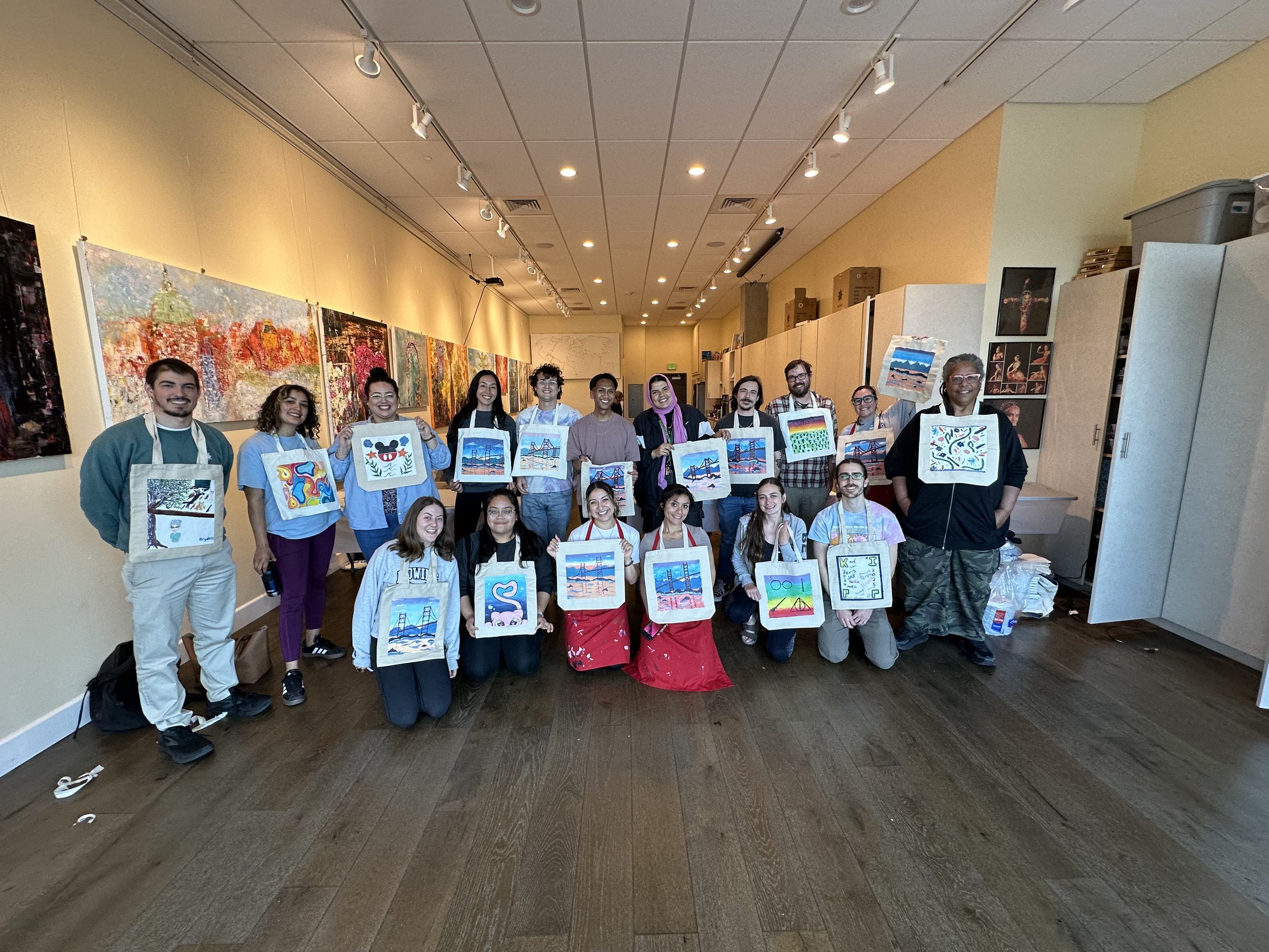 Group of people in an art studio holding up paintings they created, with colorful abstract and landscape artworks hanging on the walls.
