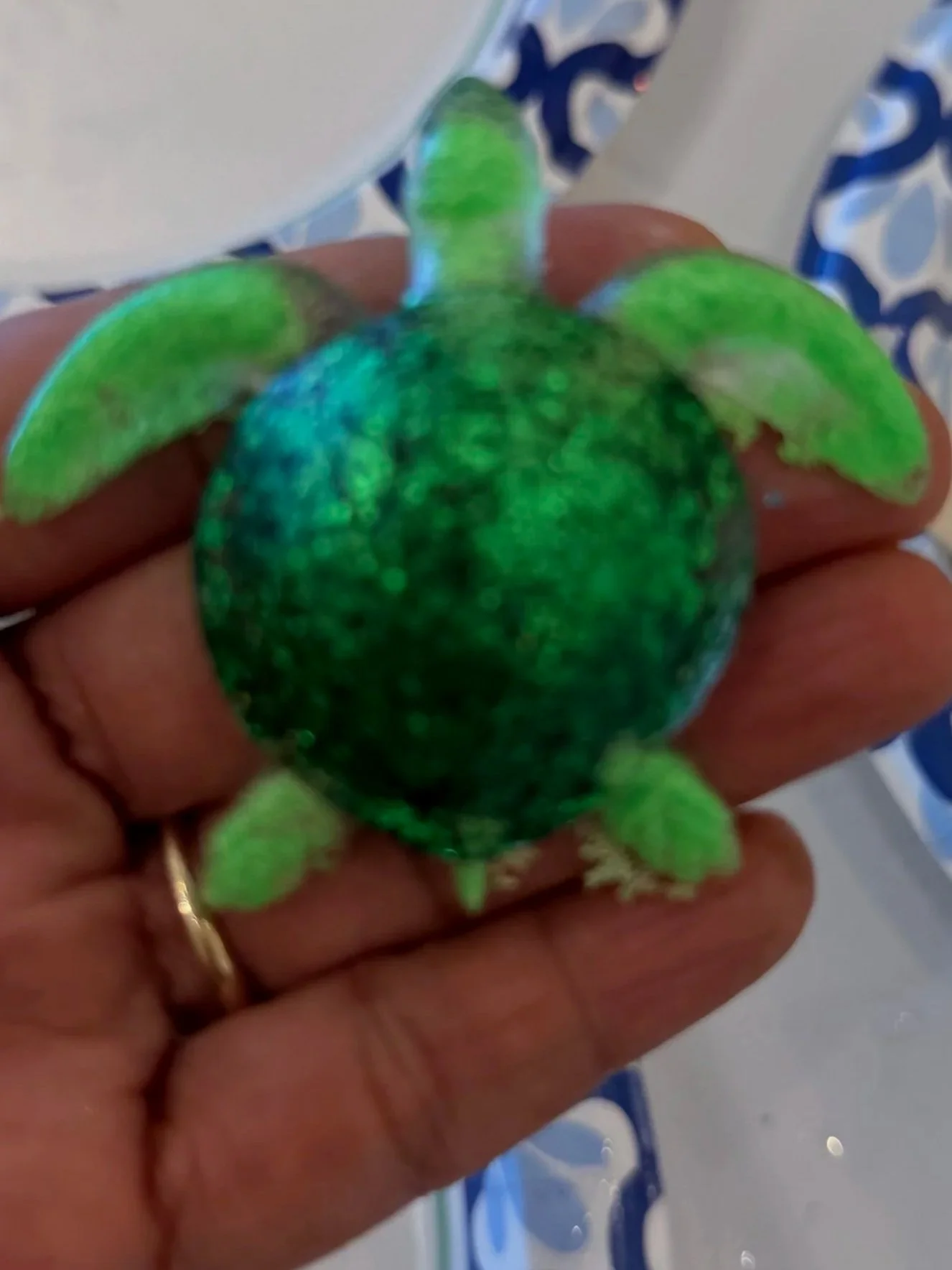 Close-up of a small toy turtle with green and blue colors being held in a person's hand. In the background, there are plates with blue and white patterns, some with pink and gold glitter, resembling decorative or craft items.