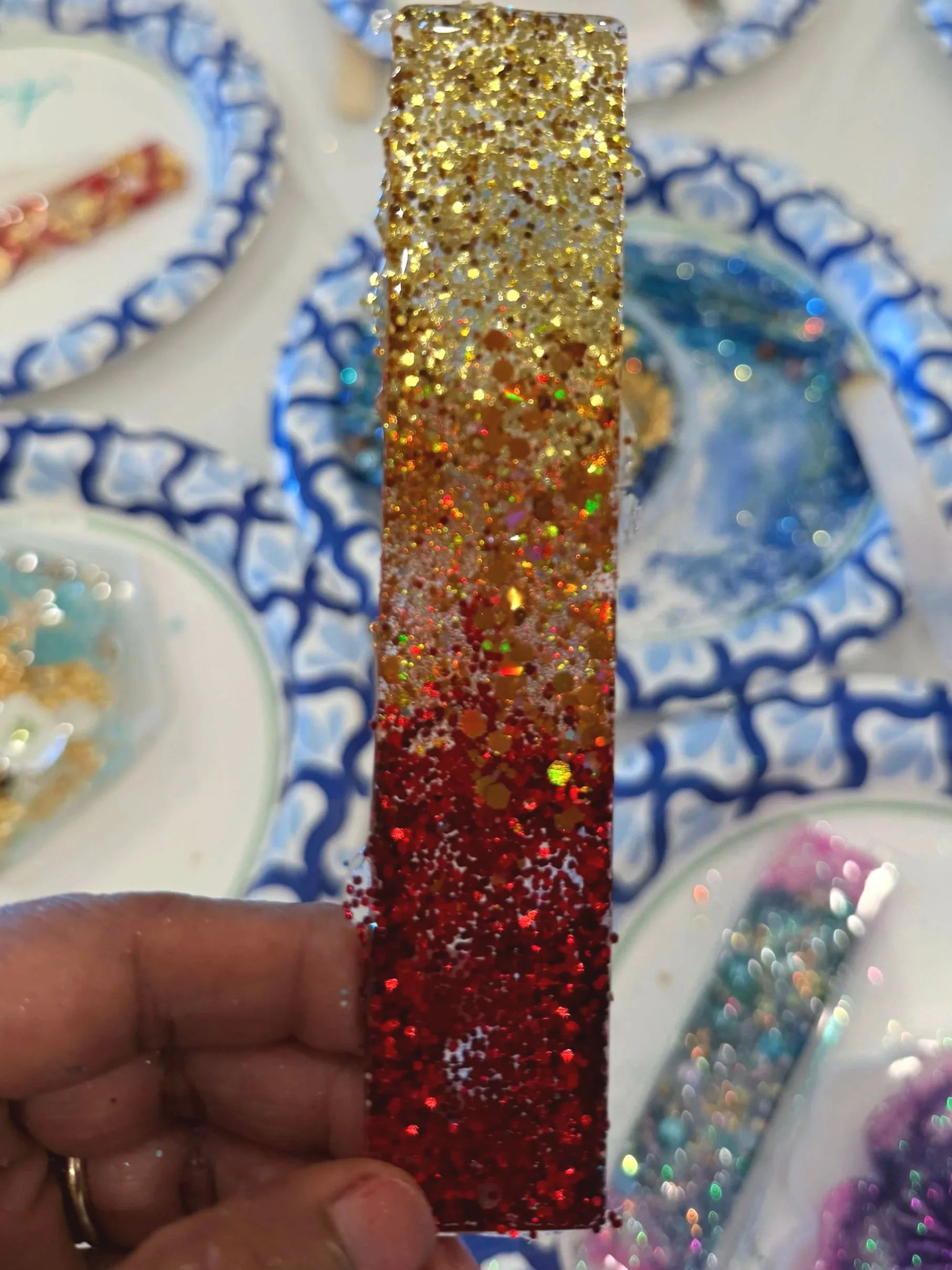 Close-up of a hand holding a rectangular piece covered in multicolored glitter, with a background of small bowls filled with various glitters and craft supplies.