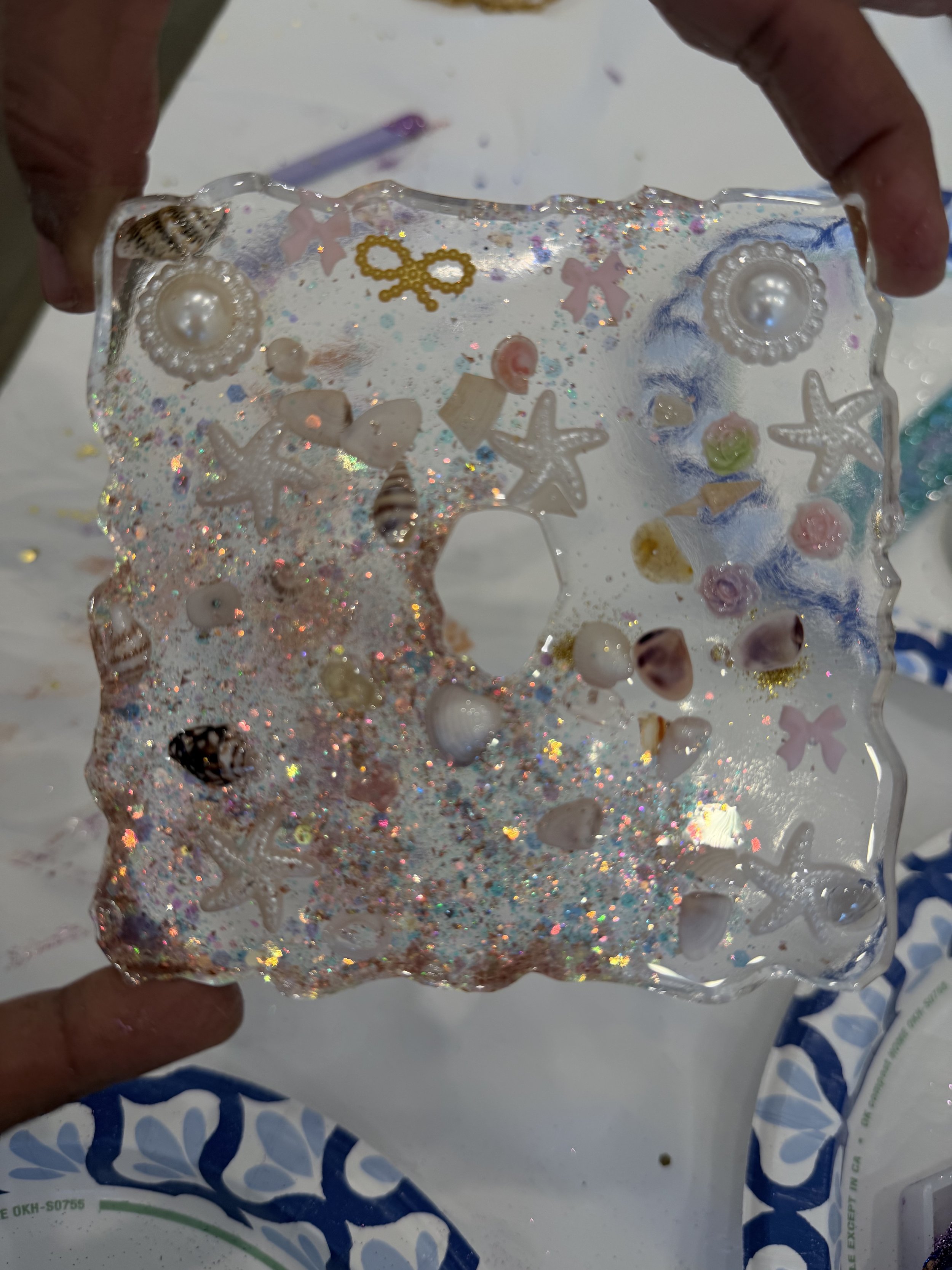 A decorative glass piece with starfish, shells, pearls, and glitter, held by a person's hands over a paper bag.