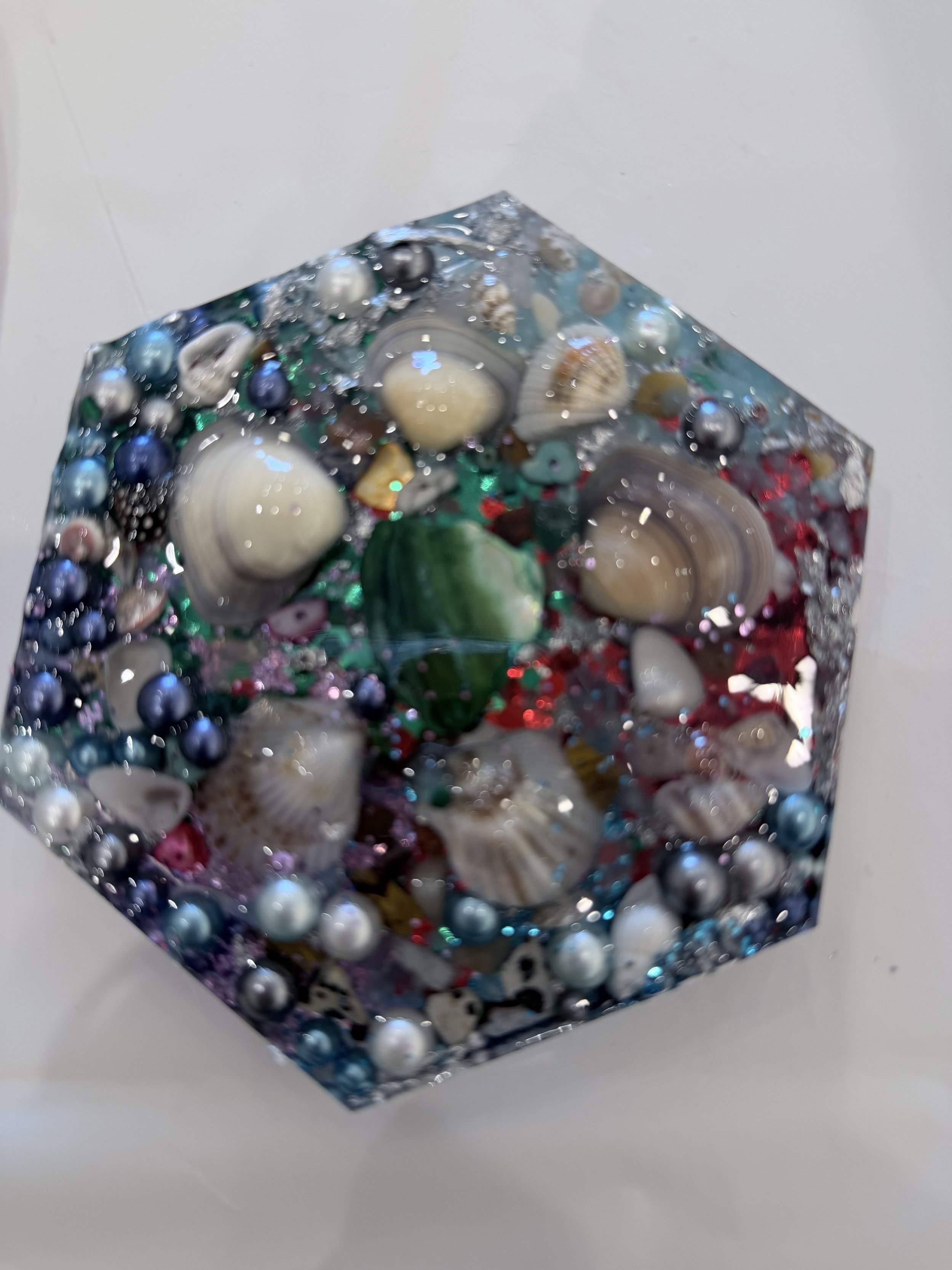 A clear rectangular block embedded with various small seashells, beads, and colorful decorative elements, placed on a white surface.