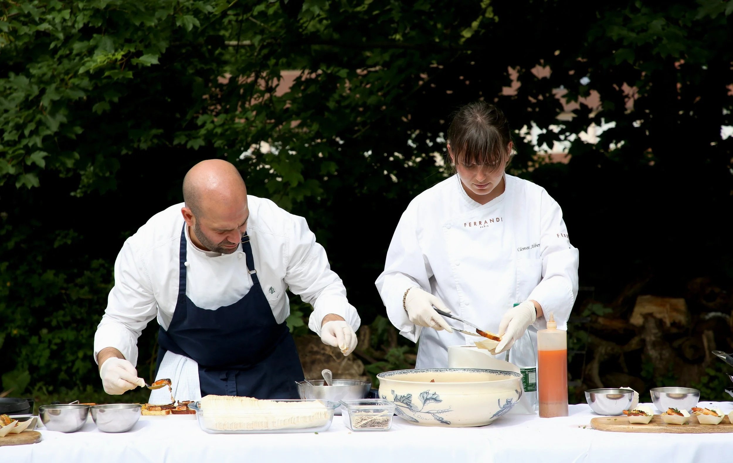 Two chefs, a man and a woman, preparing food outdoors beside a table with various bowls and ingredients, surrounded by greenery.