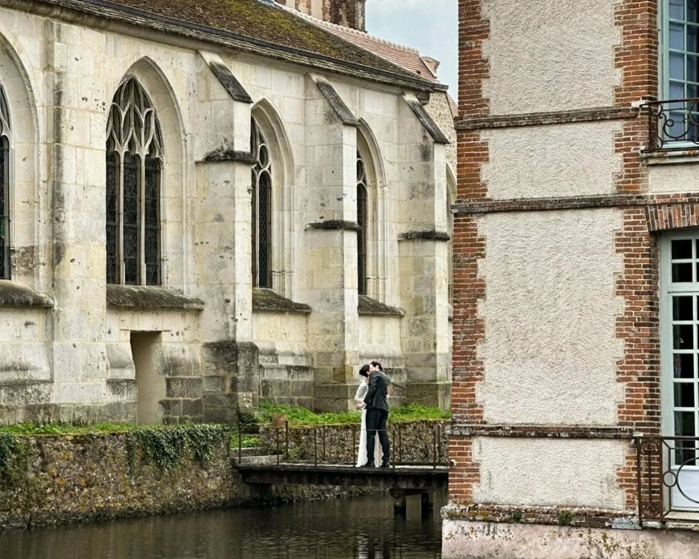 A bride and groom kiss on a small balcony overlooking a canal, with historic stone and brick buildings in the background.