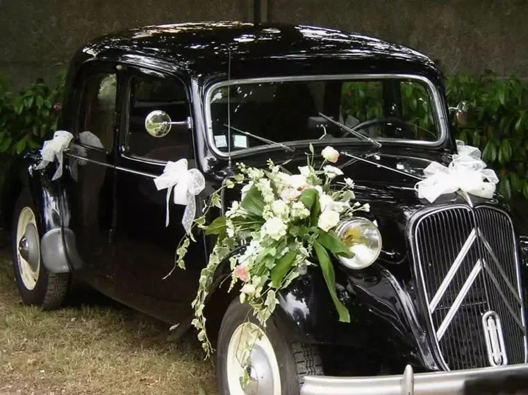 Close-up of a vintage black car with shiny chrome details, parked outdoors during daytime. The front left headlight and part of the hood are visible, with reflections of trees and sky on the car's surface.