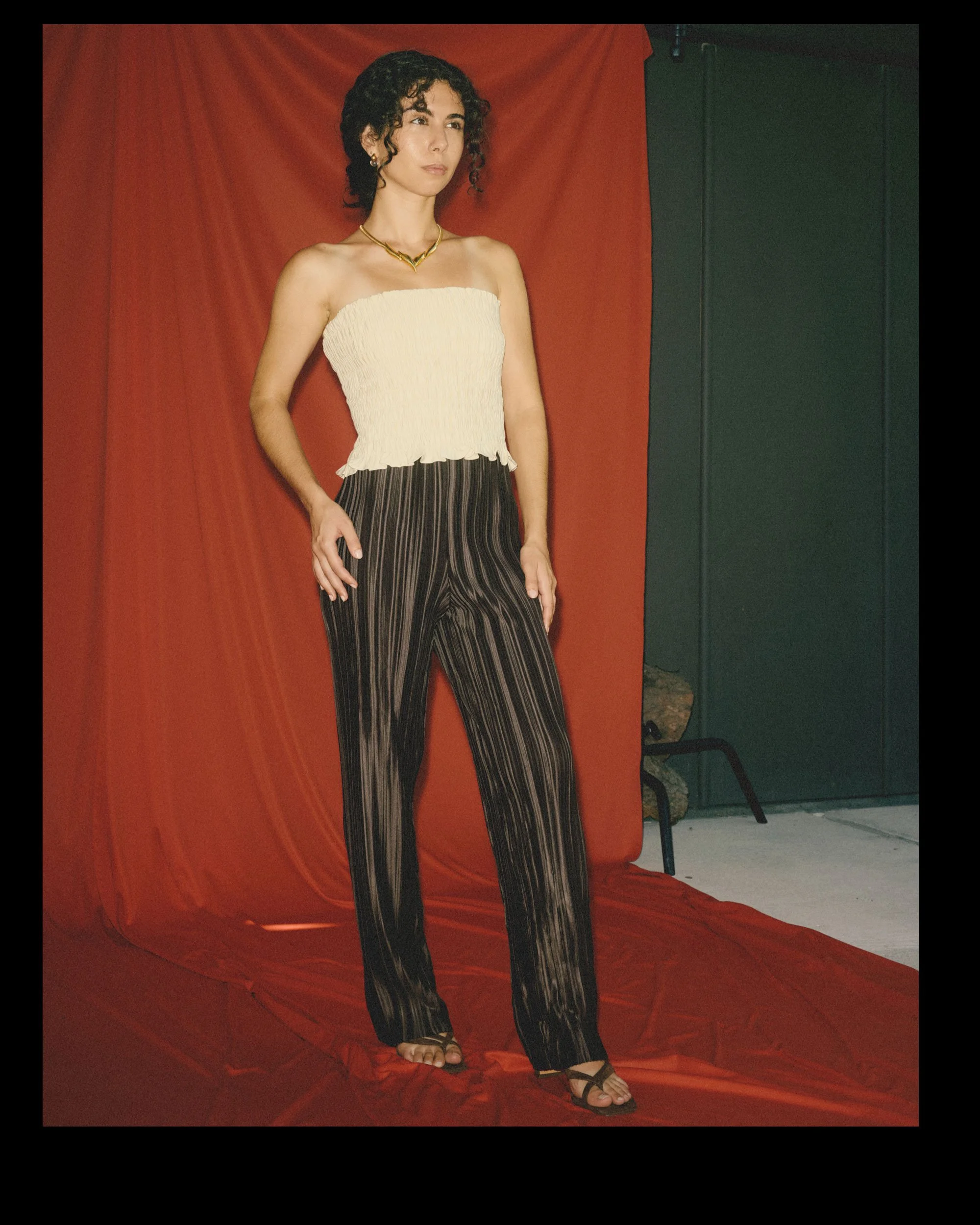 A short woman with a proportionally long torso and short legs pairs a white strapless top with horizontal shirring with full-length brown plissé pants featuring vertical folds.