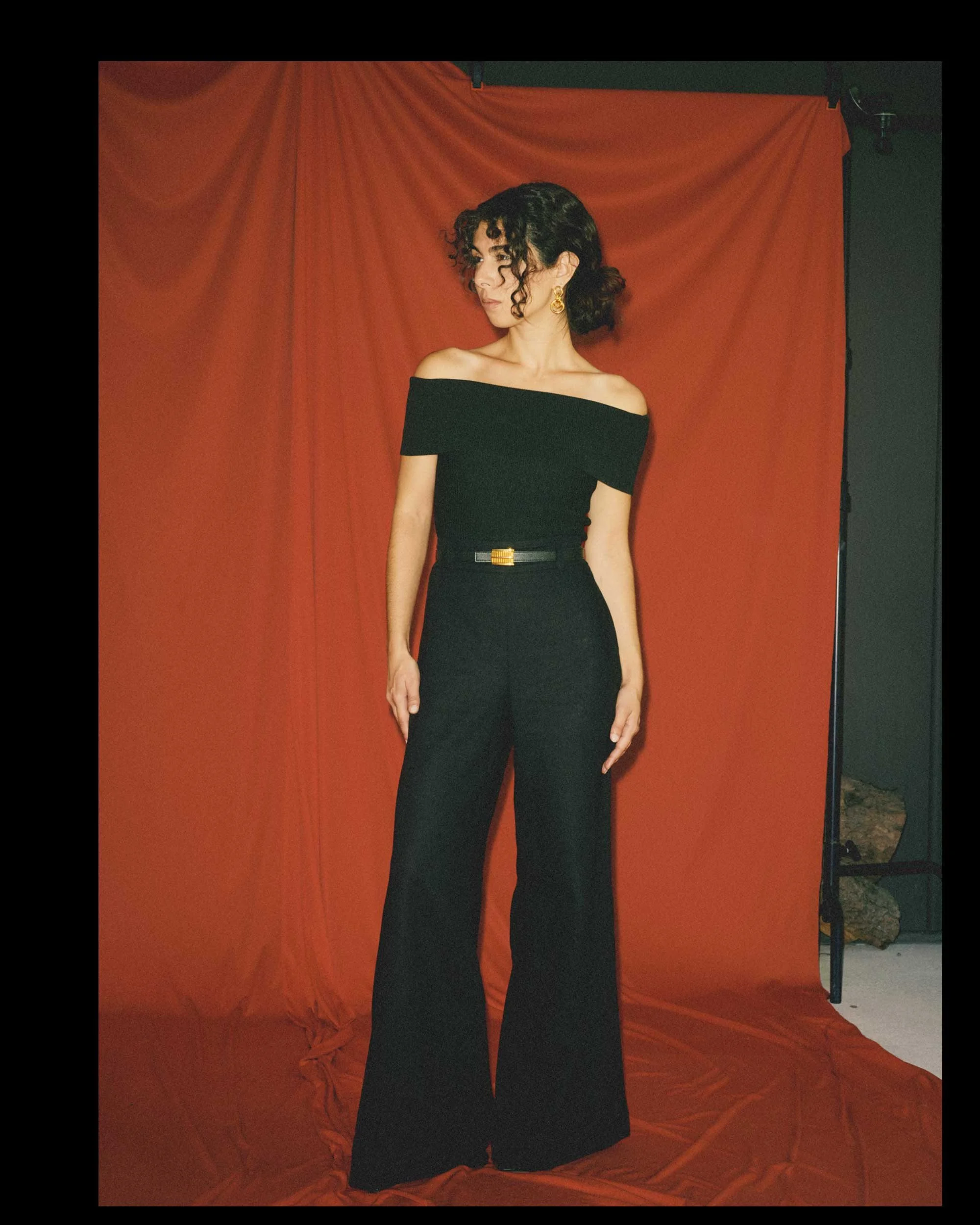 A short woman with a proportionally long torso and short legs wears a black off-the-shoulder top with high-rise, full-length palazzo pants and a black belt with a gold buckle.