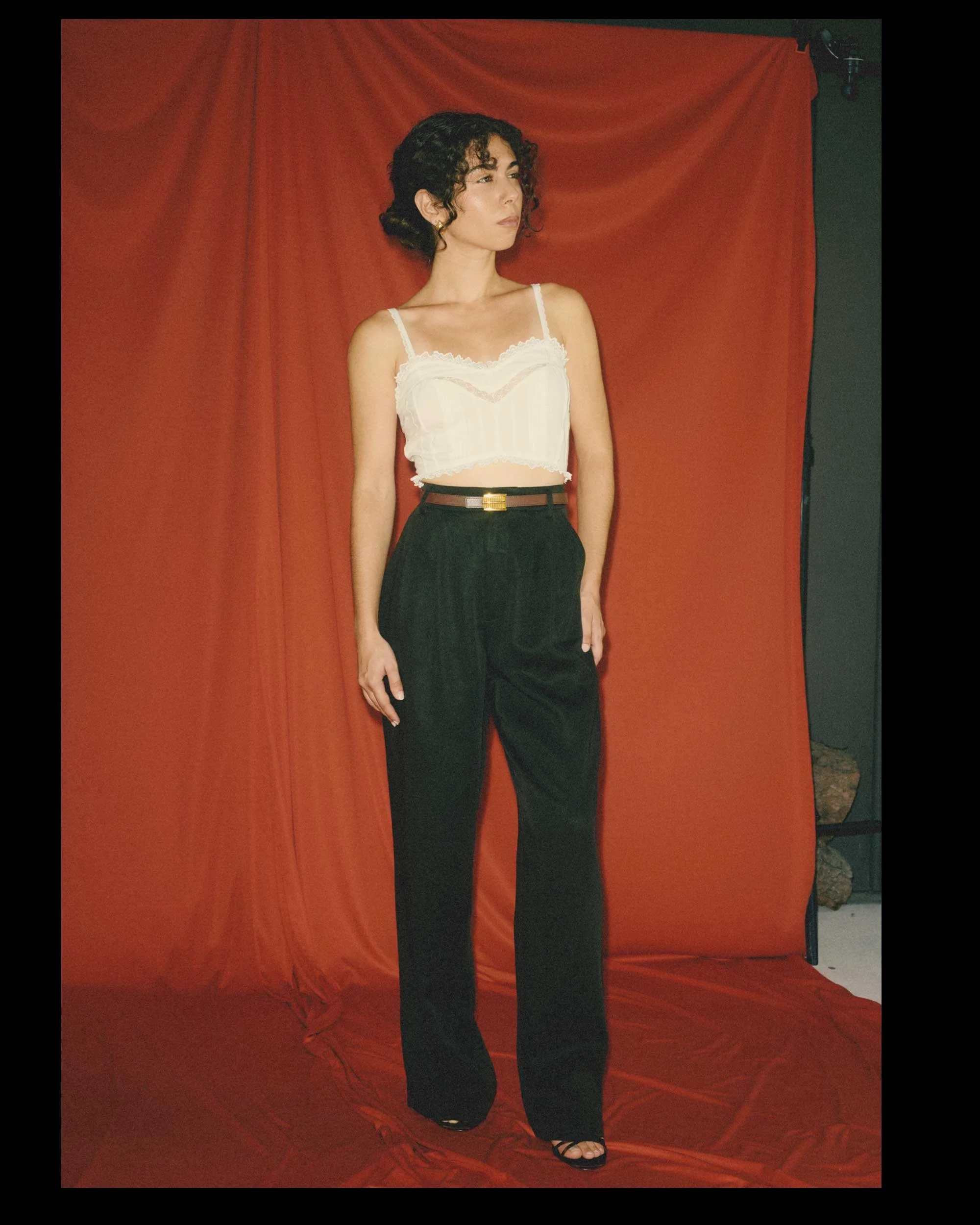 A short woman with a proportionally long torso and short legs wears a cropped white lace top with high-rise, full-length black pants and a black belt with a gold buckle.