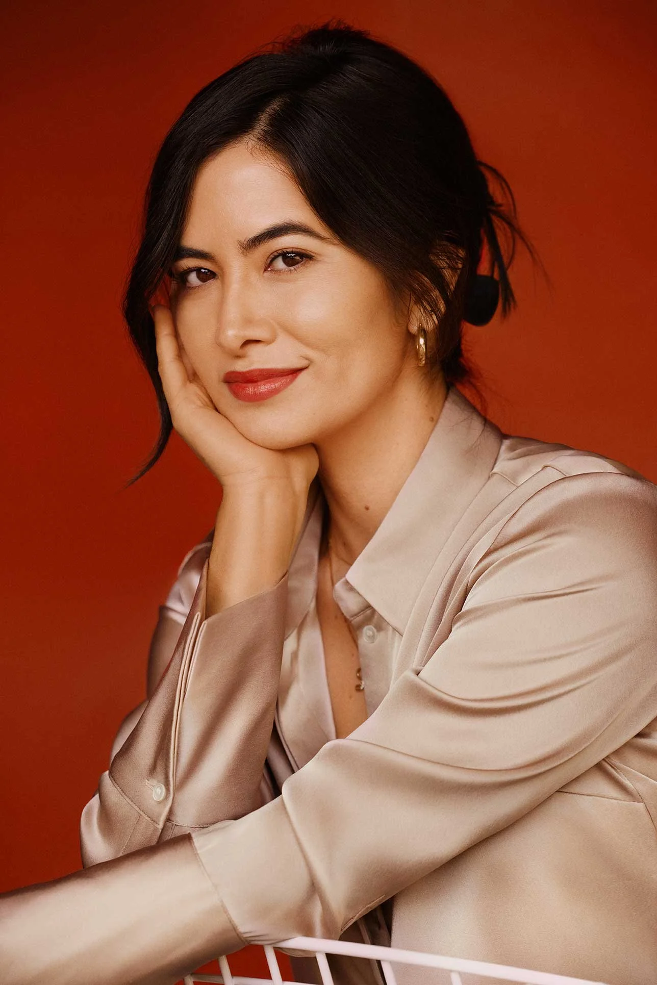 Lwany, founder of Petite Fashion Digest, wearing a champagne satin blouse with her face resting on her hand against a red backdrop.