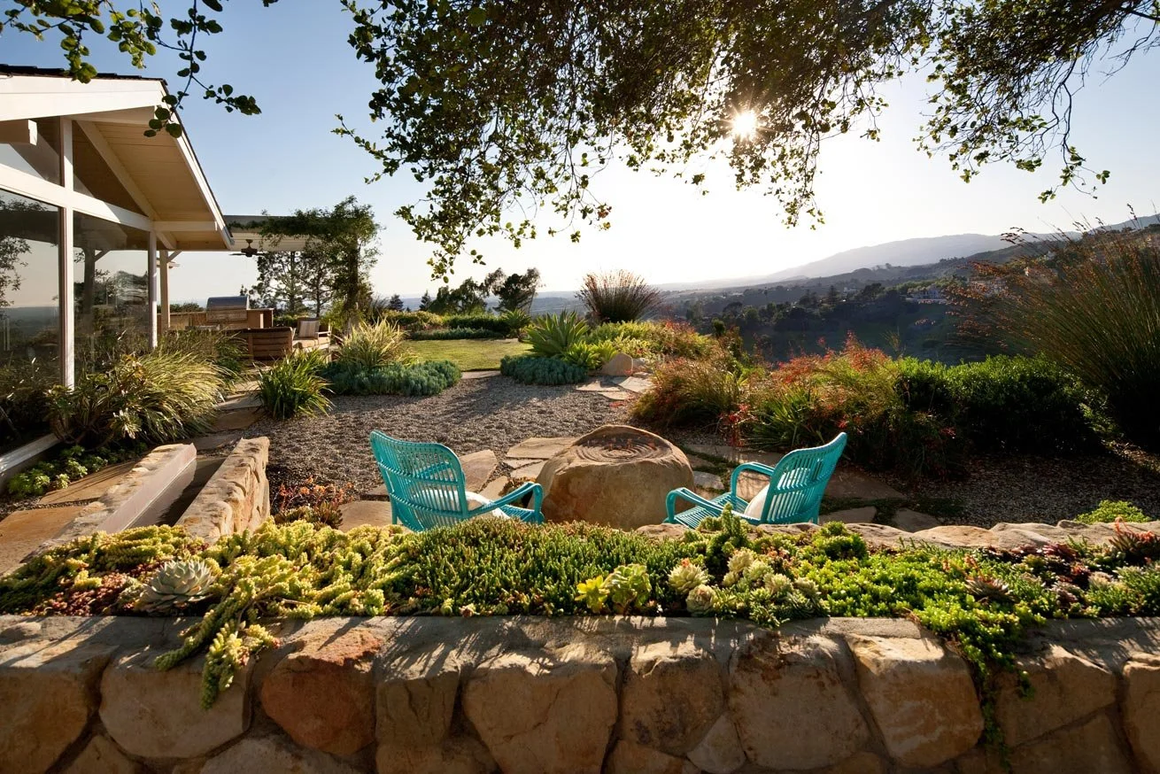 Scenic backyard with turquoise chairs overlooking a landscape view, surrounded by lush greenery, plants, and a stone patio.