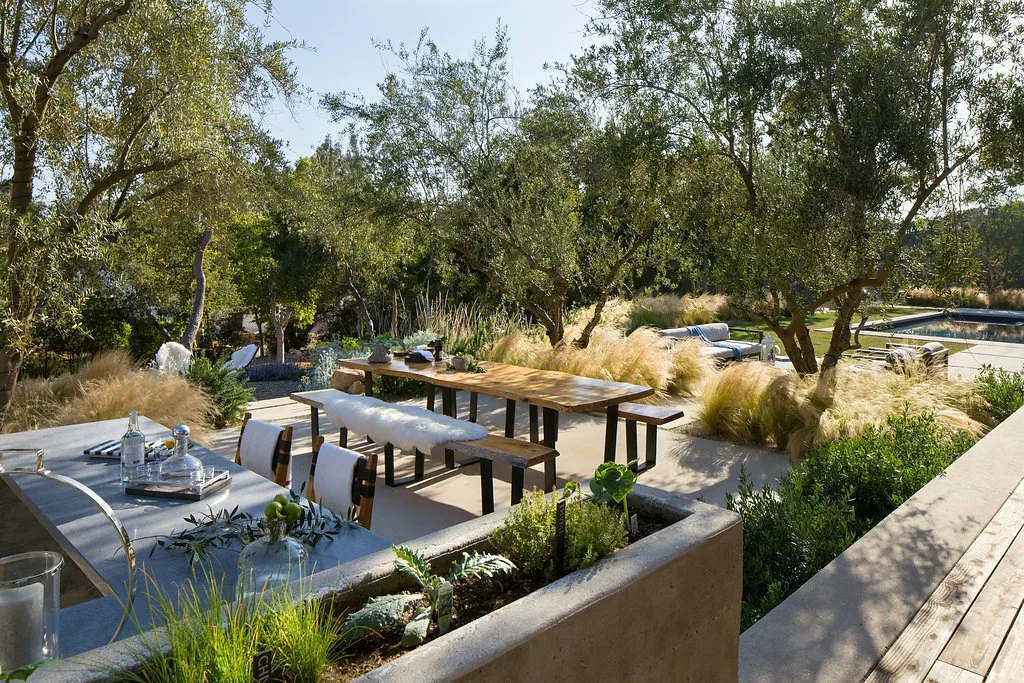 Outdoor dining area with benches and tables surrounded by lush greenery and trees, adjacent to a patio with seating and a pool.