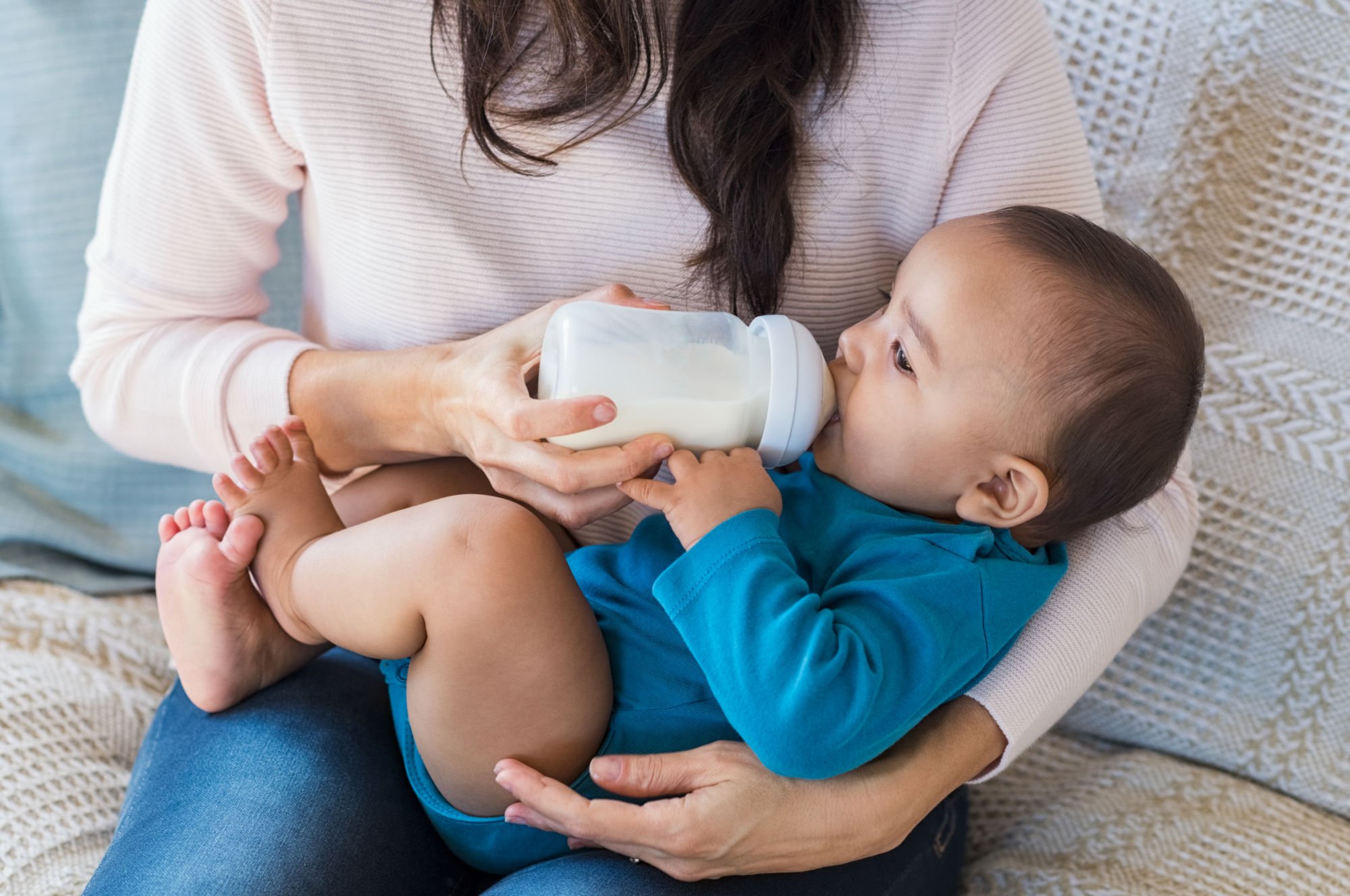 Baby wearing blue onesie being held and bottle fed by a woman wearing a pale pink shirt and jeans sitting on a sofa_PBB.jpg