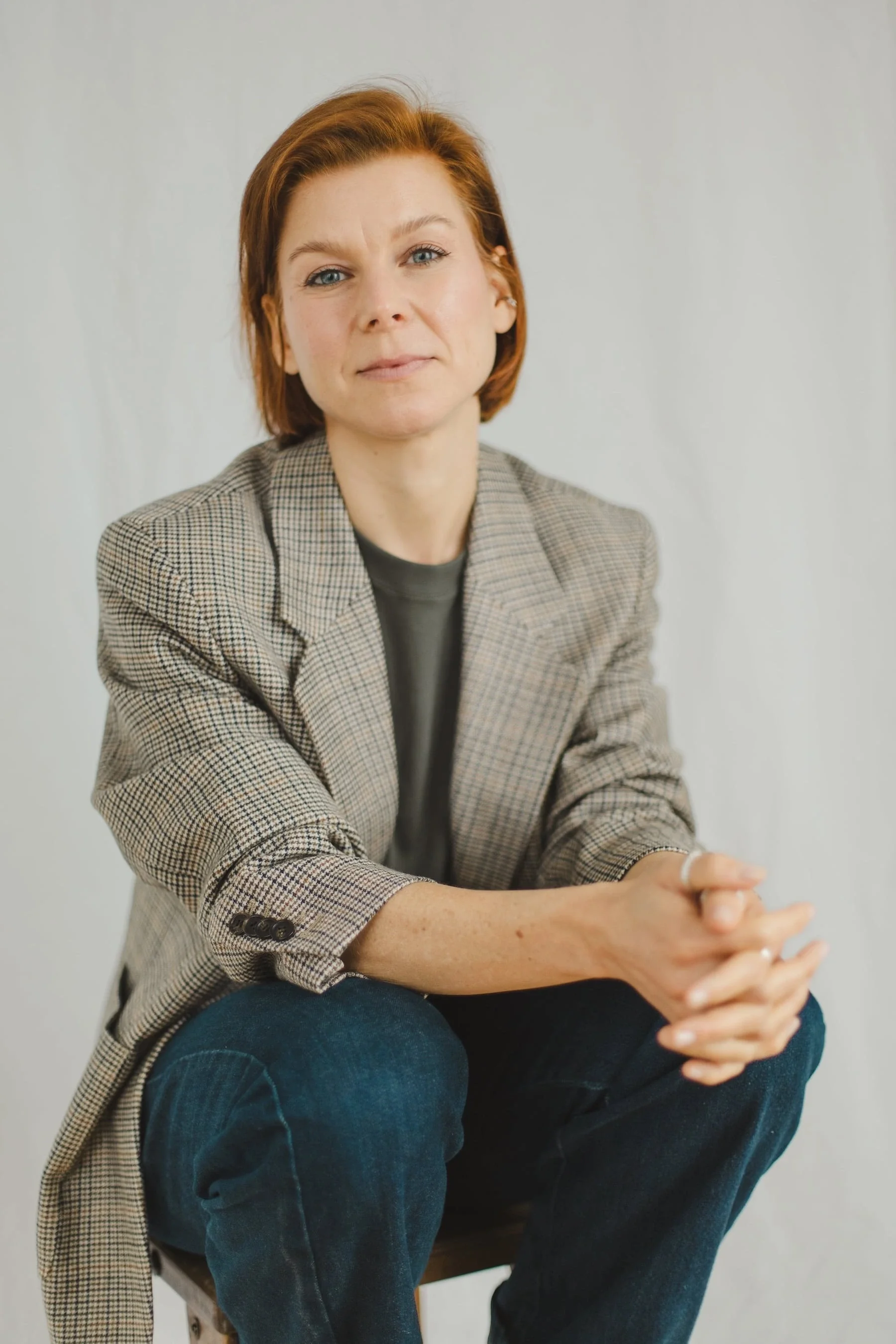 Kristina Appel mit kurzen, rötlichen Haaren, die einen karierten Blazer, ein dunkles Hemd und Jeans trägt, sitzt mit verschränkten Händen da und blickt selbstbewusst in die Kamera.