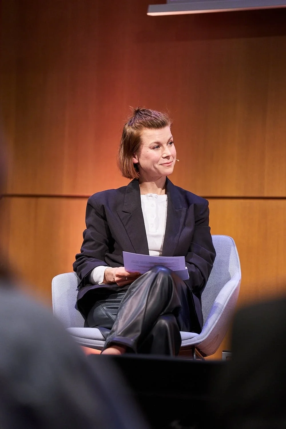 Moderatorin Kristina Appel sitzt auf einer Bühne auf einem Stuhl, hält Moderationskarten in der Hand und schaut aufmerksam zur Seite. Sie trägt einen dunklen Blazer über einem weißen Hemd und einer dunklen Hose.