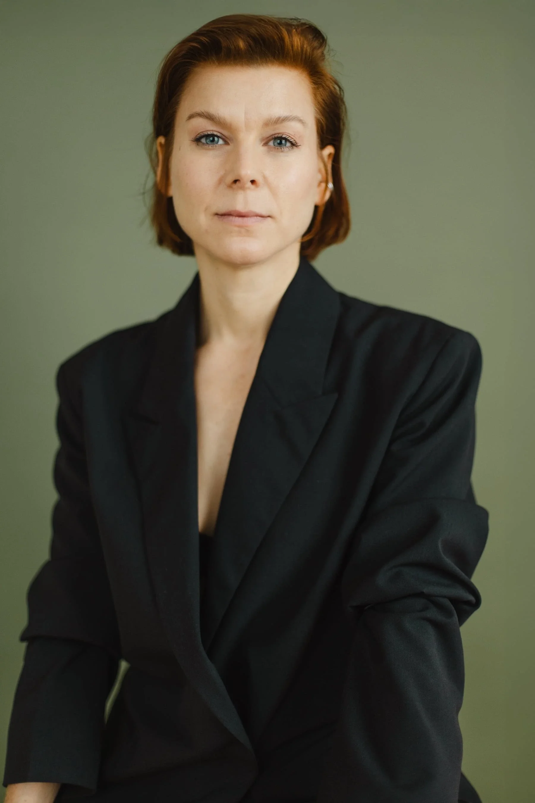 Kristina Appel, mit kurzem, rotbraunem Haar trägt einen schwarzen Blazer und sitzt vor einem schlichten, gedeckten grünen Hintergrund, wobei sie mit neutralem Gesichtsausdruck direkt in die Kamera blickt.