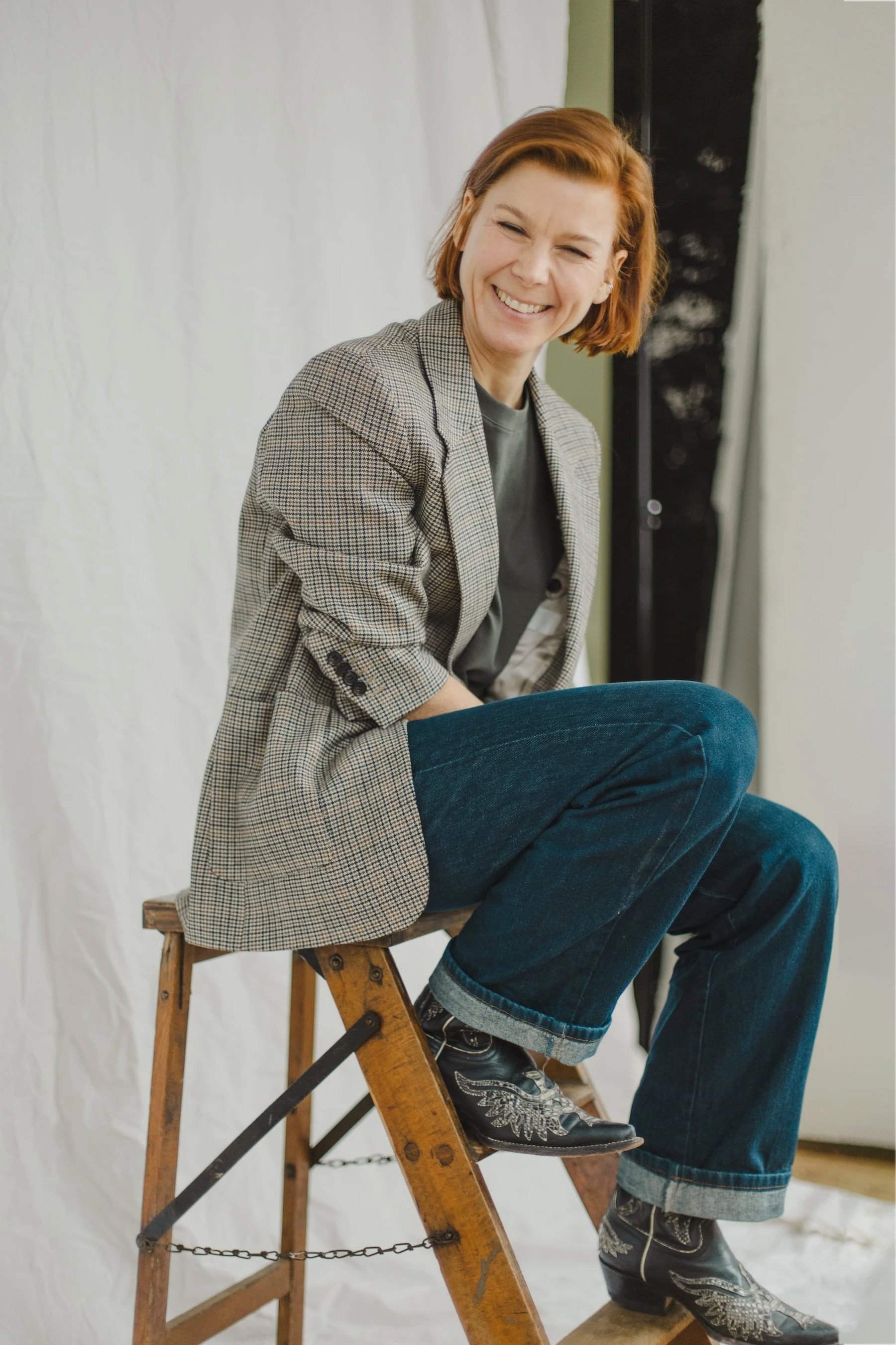 Kristina Appel, mit kurzen, rotbraunen Haaren sitzt auf einem hölzernen Tritthocker und lächelt. Sie trägt einen karierten Blazer, ein dunkles Hemd, eine Jeans mit weitem Bein und bestickte Cowboystiefel.