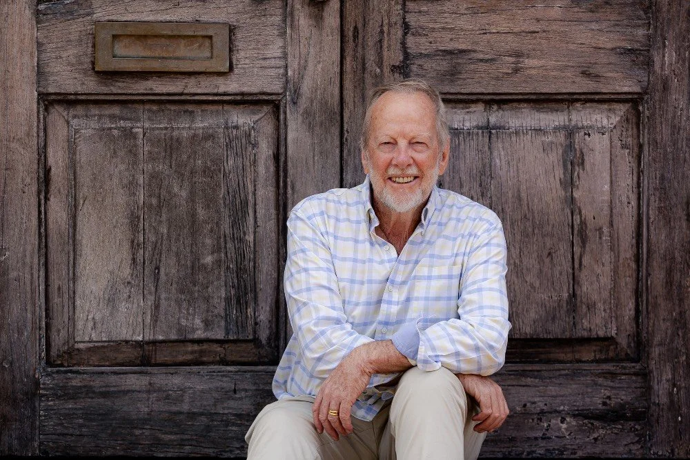 Elderly man with beard, collared shirt and cream pants, sitting in front of a weathered wooden door photographed by Kirsten Graham Photography