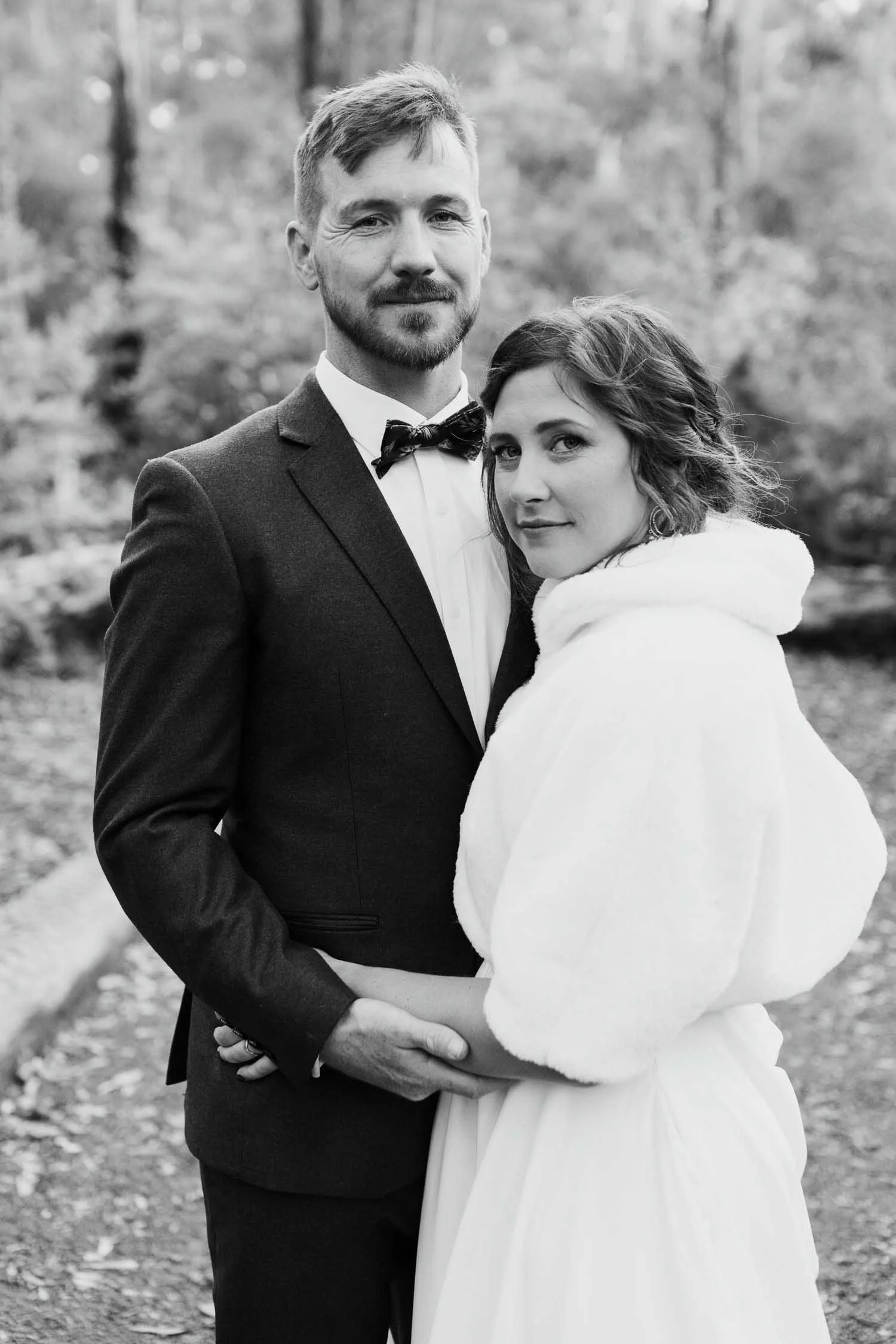 Bride and groom standing outdoors, looking at camera, photographed in black and white by Clements + Co. Studio Photography.