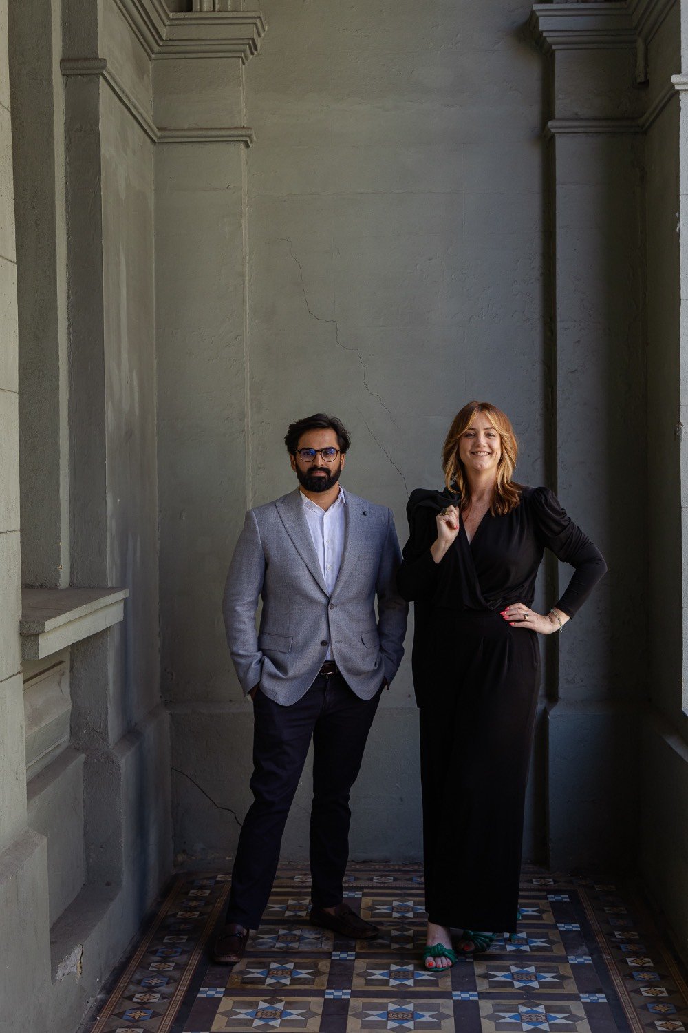 Man with beard, light grey blazer and navy pants standing next to a red haired lady in a black dress, in a heritage building foyer in Fremantle, corporate photography by Kirsten Graham Photography.