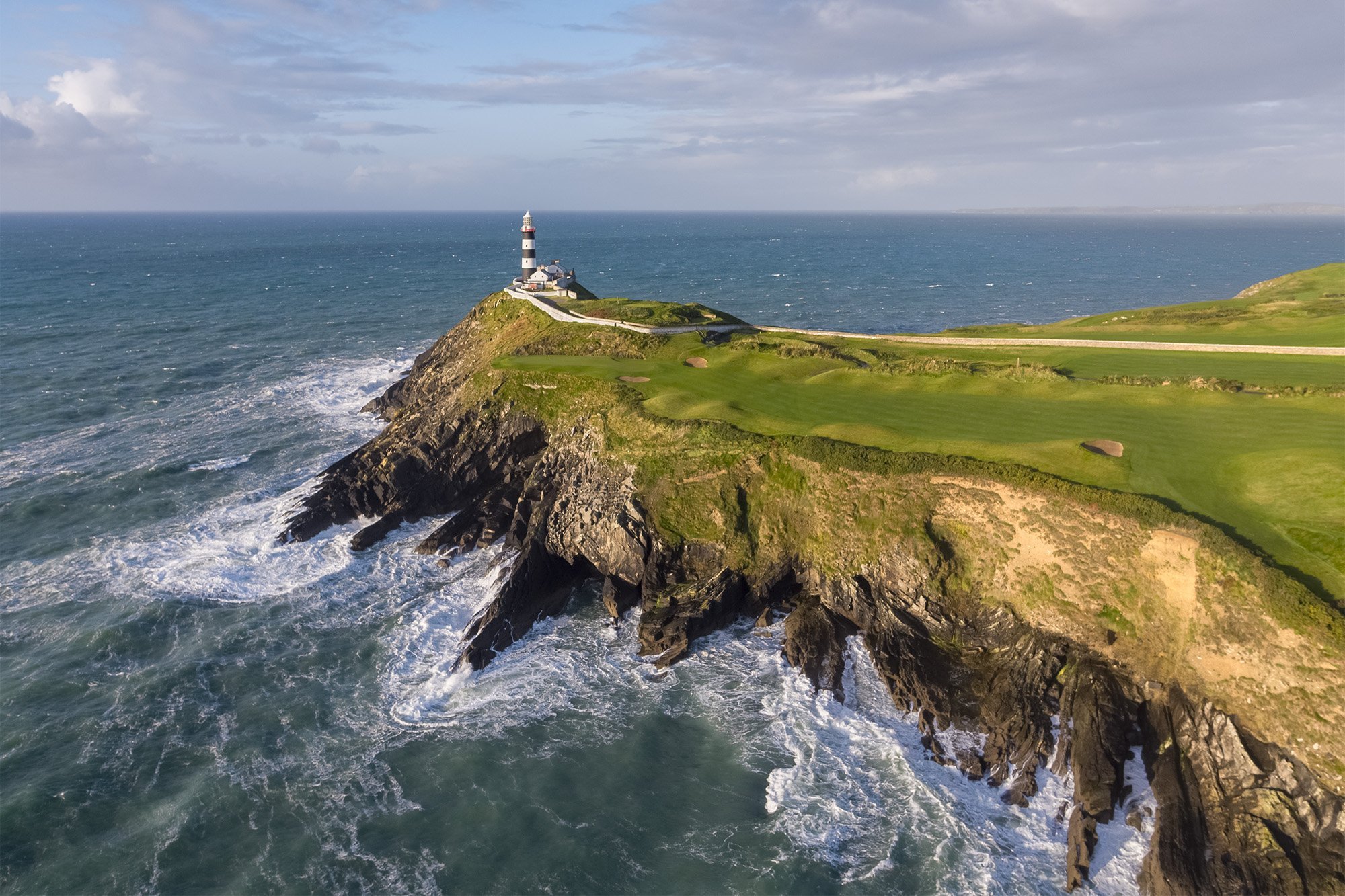 A coastal landscape with a lighthouse on a cliff overlooking the ocean, with waves crashing against the rocks below, and a green grassy area around the lighthouse.