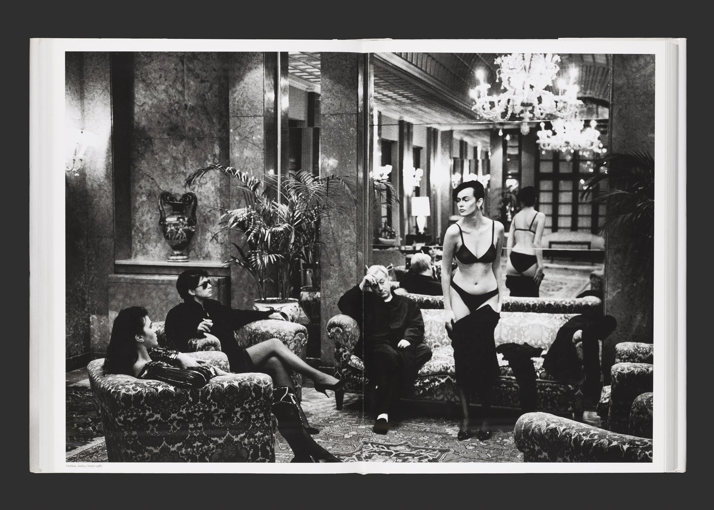 Une scène en noir et blanc dans un hall luxueux avec des personages dont deux femmes en lingerie, une assise et une debout, ainsi que deux autres femmes en lingerie en arrière-plan.