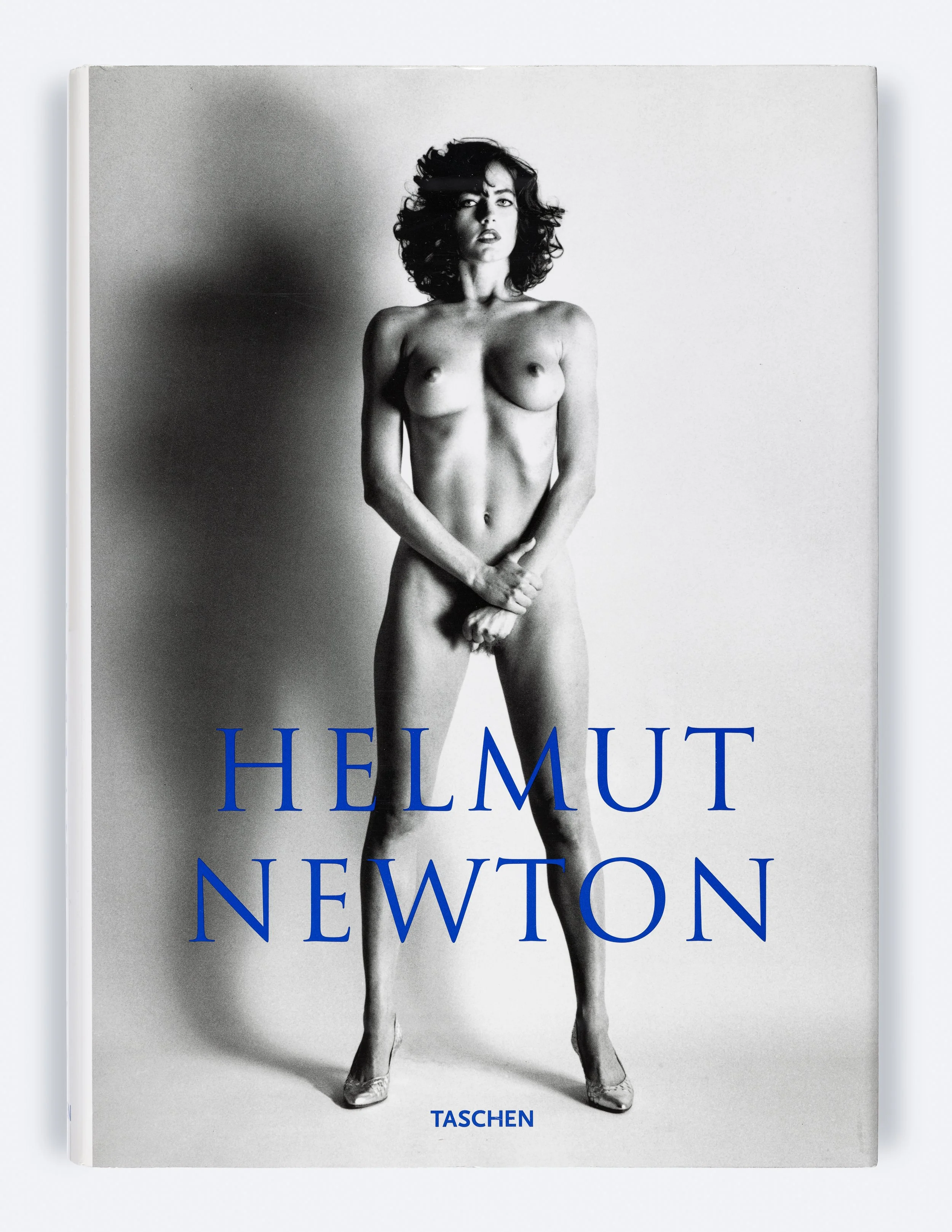 Couverture d'un livre avec une photographique en noir et blanc d'une femme nue avec les jambes écartées et les mains devant la région génitale, le tout avec le titre "Helmut Newton" en bleu et le nom du éditeur "Taschen" en bas.