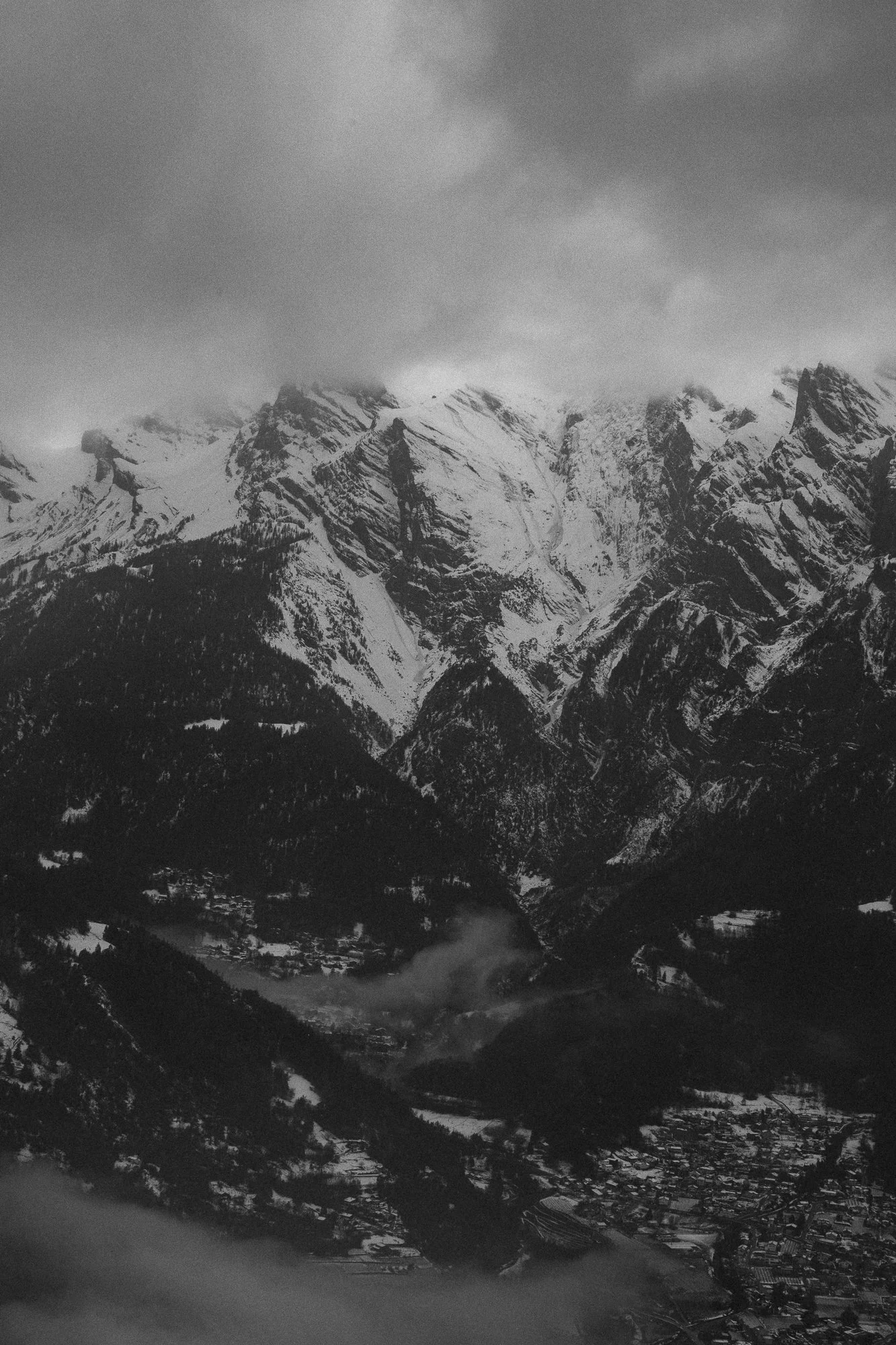 Montagnes enneigées sous un ciel nuageux, vue d'ensemble d'une vallée alpine avec village, arbres et nuages dans une image en noir et blanc.