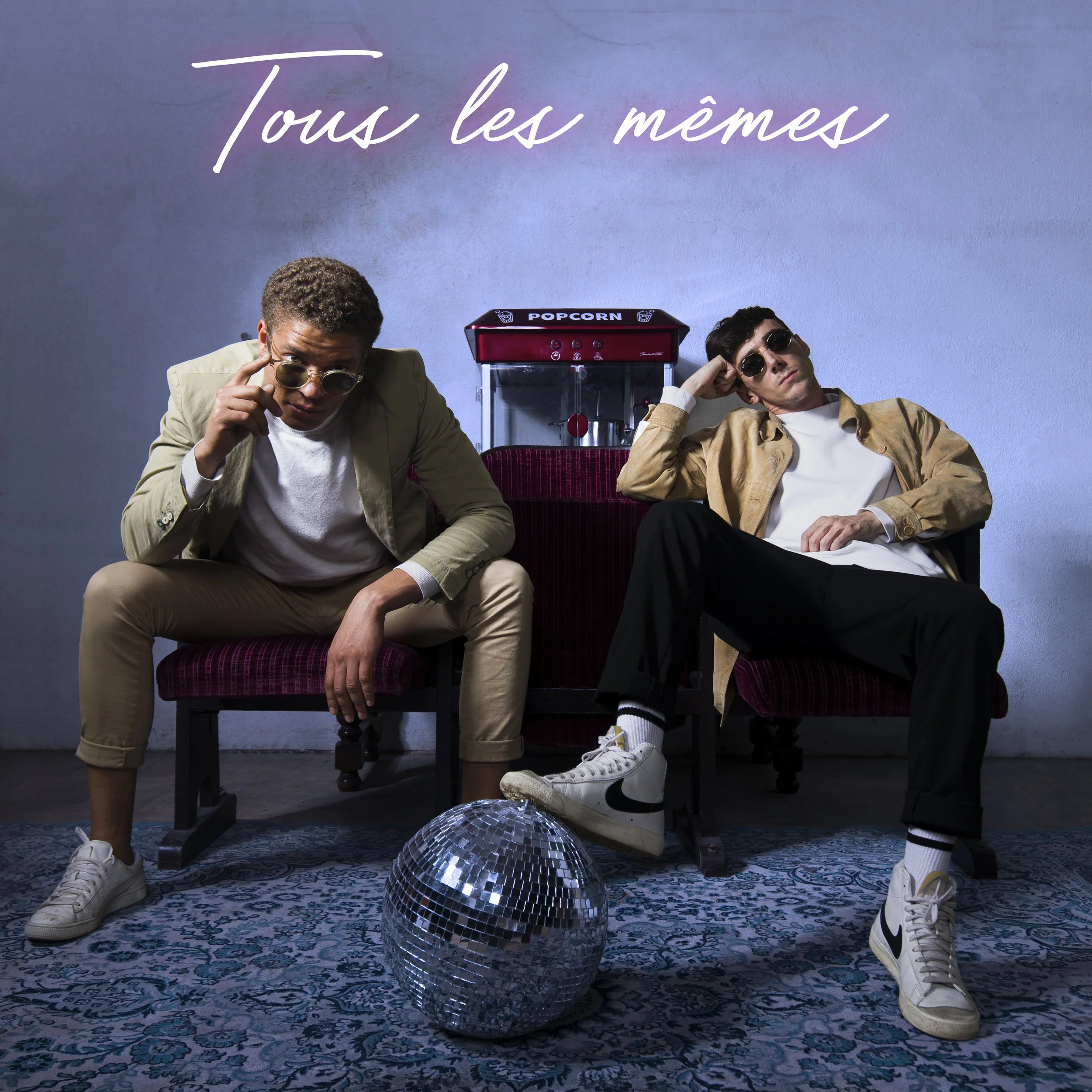 Deux hommes assis dans une pièce avec un tapis bleu et une boule disco devant eux. L'un porte un costume beige, l'autre un manteau beige, tous deux avec des lunettes de soleil. Il y a un distributeur de popcorn en arrière-plan et le texte en haut dit