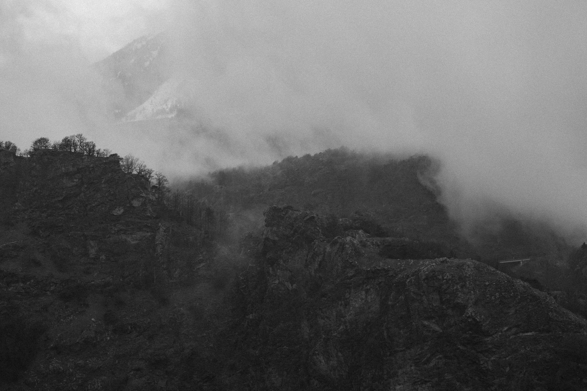 Paysage montagneux brumeux en noir et blanc avec des rochers et des arbres dispersés.