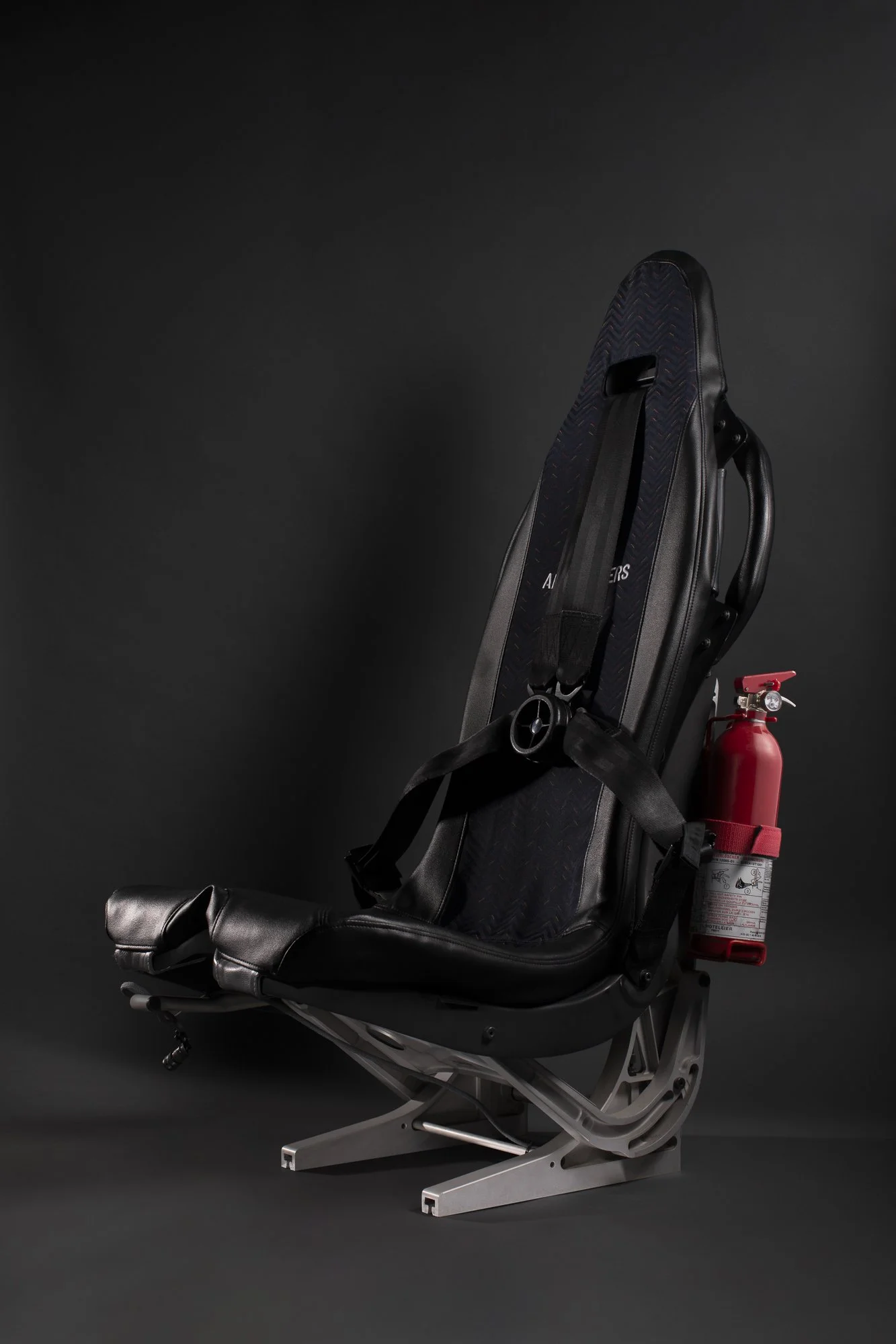 Une chaise d'aéroport avec un extincteur attaché à l'arrière. La chaise est noire avec un coussin intégré, sur un fond noir