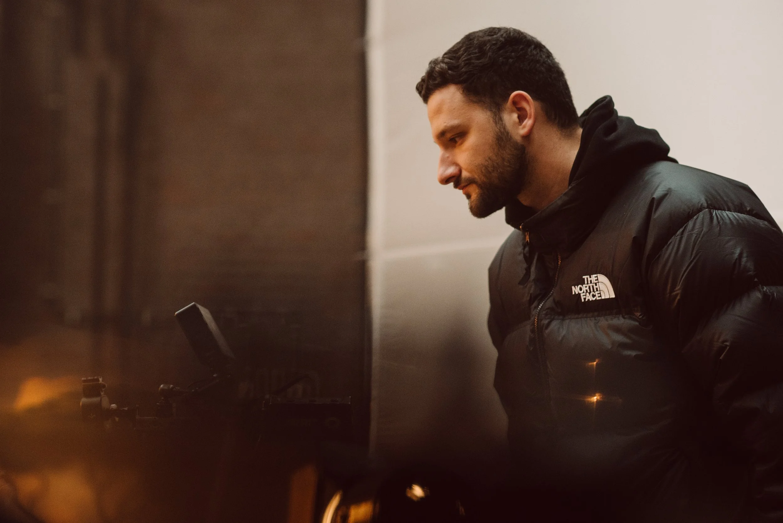 A man with dark hair and a beard wearing a black North Face jacket, leaning forward and working on a piano. director commercial 