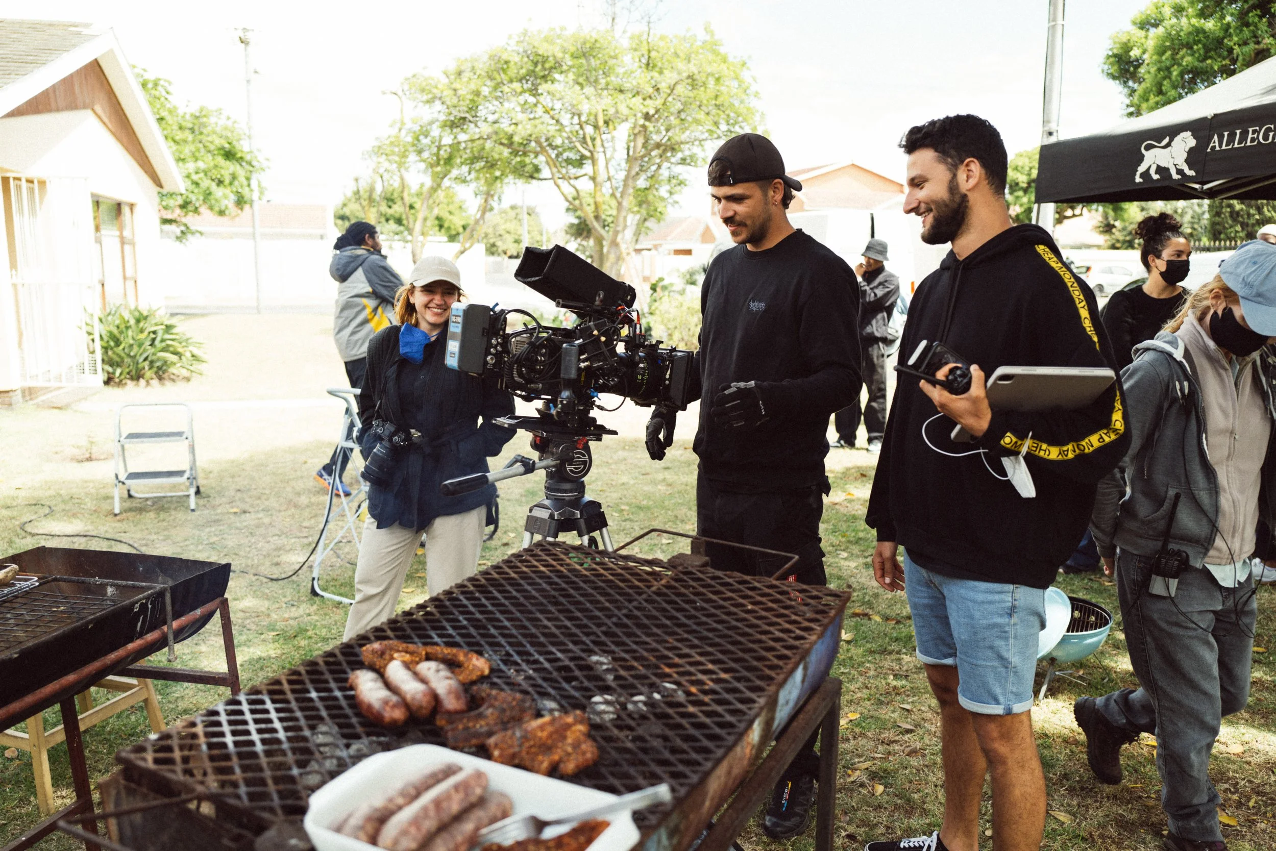 Group of people filming at an outdoor BBQ setup, with a professional camera and grilled sausages in the foreground.