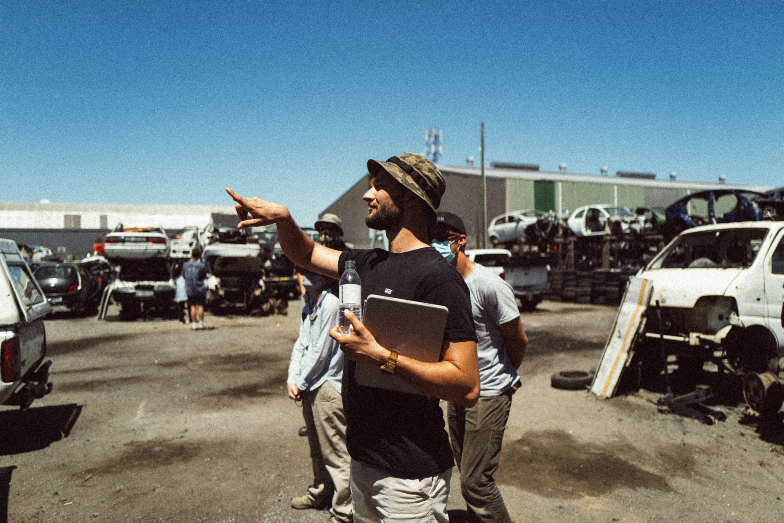 Group of people in a junkyard with stacked cars, one person pointing, holding a bottle and a laptop, wearing a camo bucket hat.