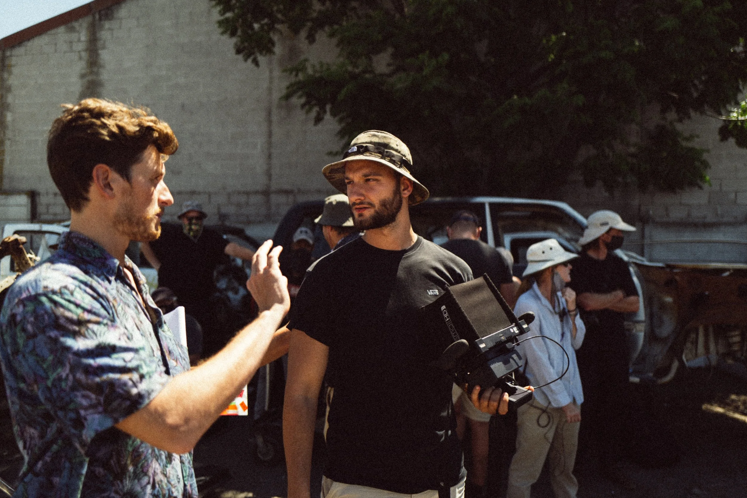 Two men in casual attire, one holding a camera, engaged in a discussion outdoors with several people in the background wearing hats and standing near vehicles.