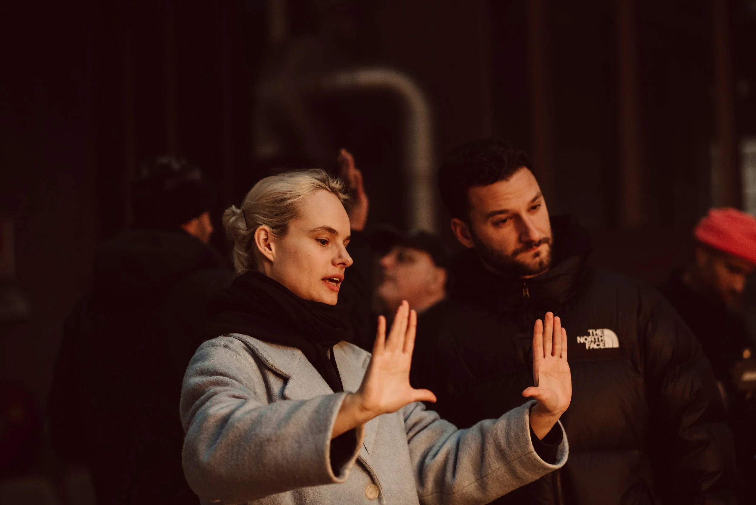 A woman with blonde hair and a man with dark hair and a beard are standing outside. The woman is holding her hands up as if blocking or protesting, and the man is also holding his hands up with palms facing outward. They are dressed warmly in jackets