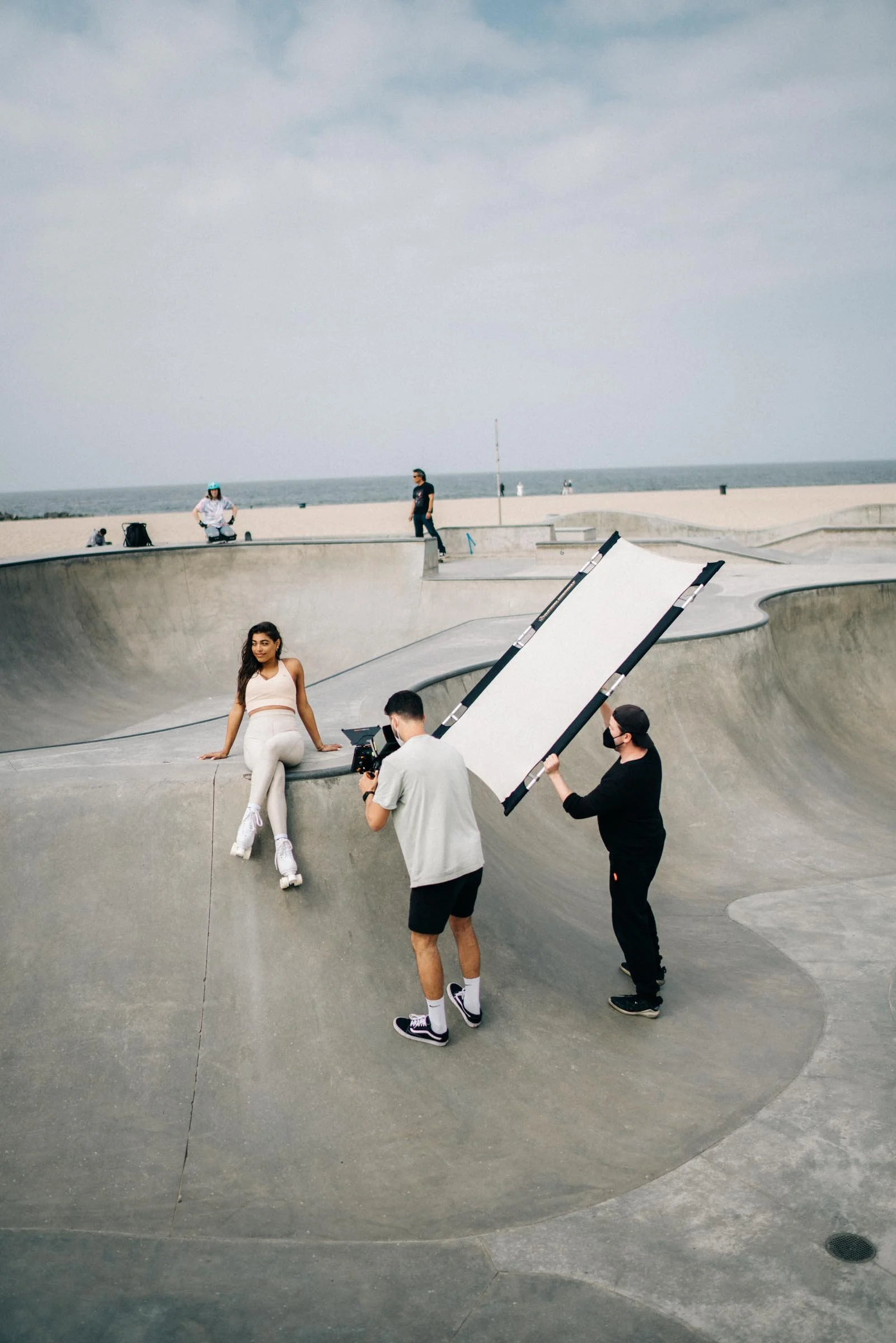A woman in roller skates posing at a skatepark while two men with equipment film her. The scene is set on a concrete ramp with a beach and ocean in the background.