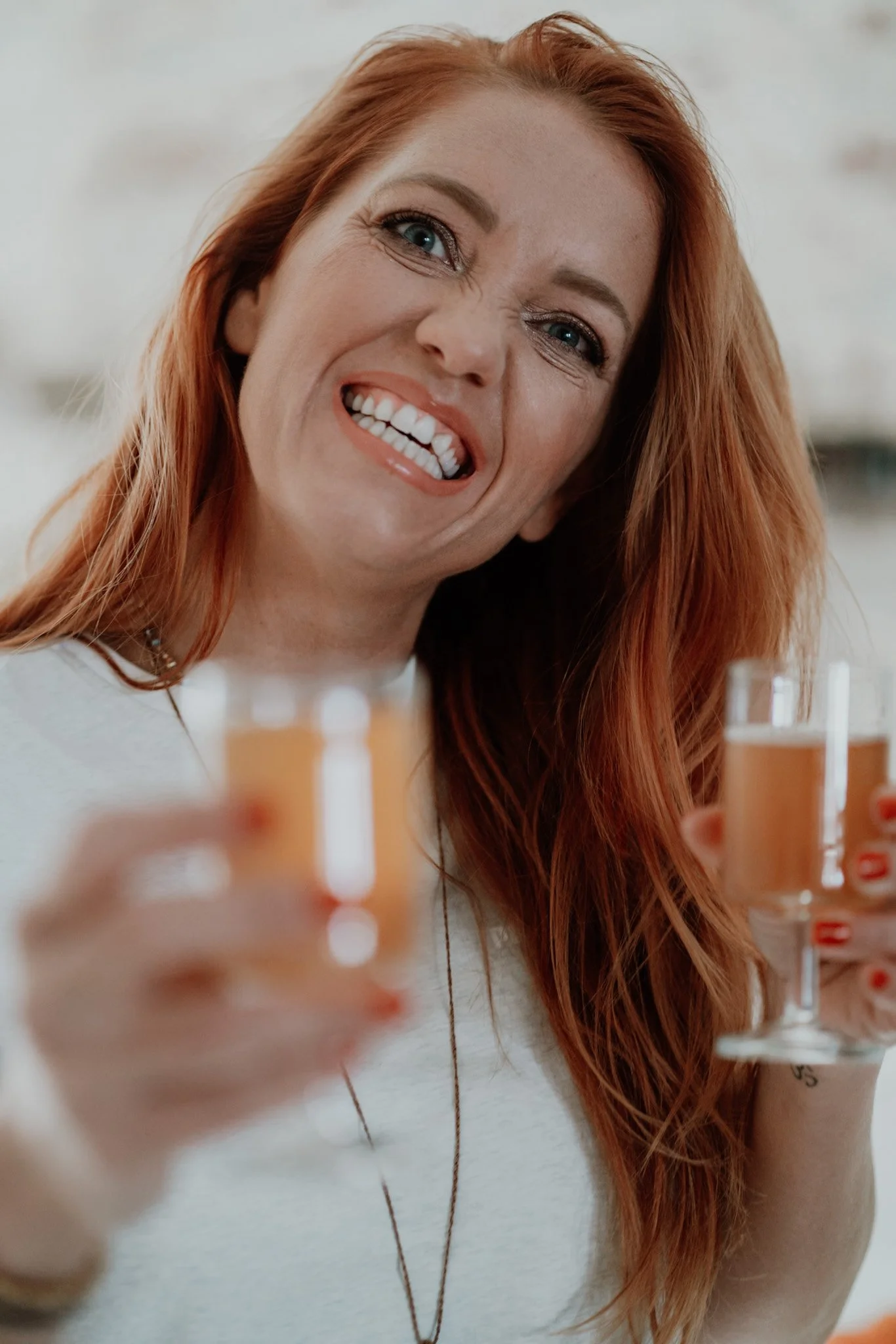 A red-haired woman making a funny face while holding two glasses of orange-colored drinks.