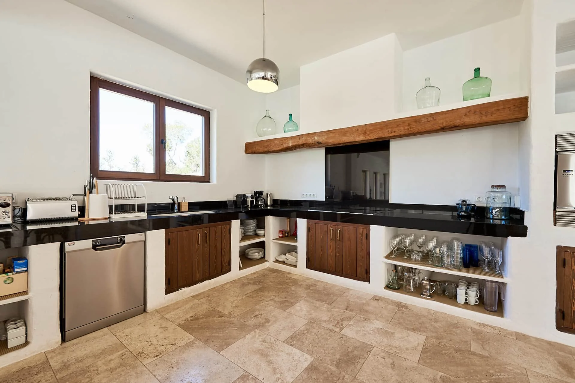 Kitchen with black countertops, brown wooden cabinets, a large window, and glass bottles on a wooden shelf above the firebox.
