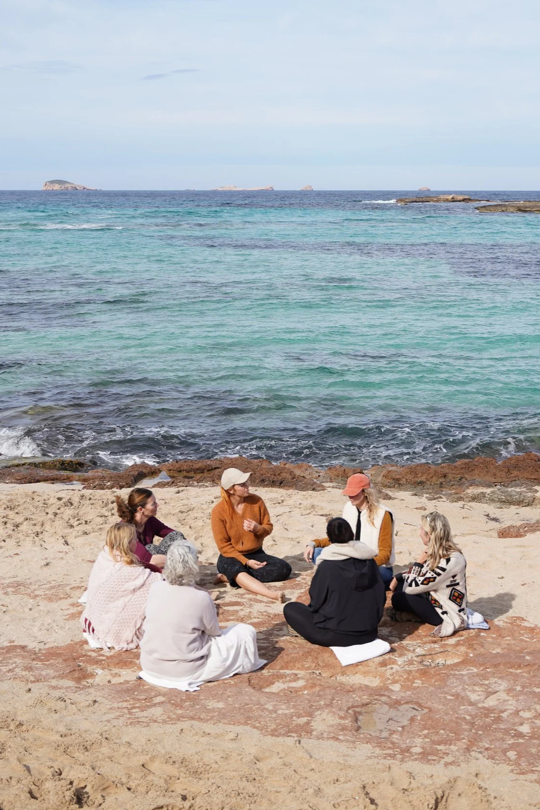 Group of seven people sitting and conversing on a sandy beach near the ocean, with rocks along the shoreline and small islands visible in the distance under a slightly cloudy sky.