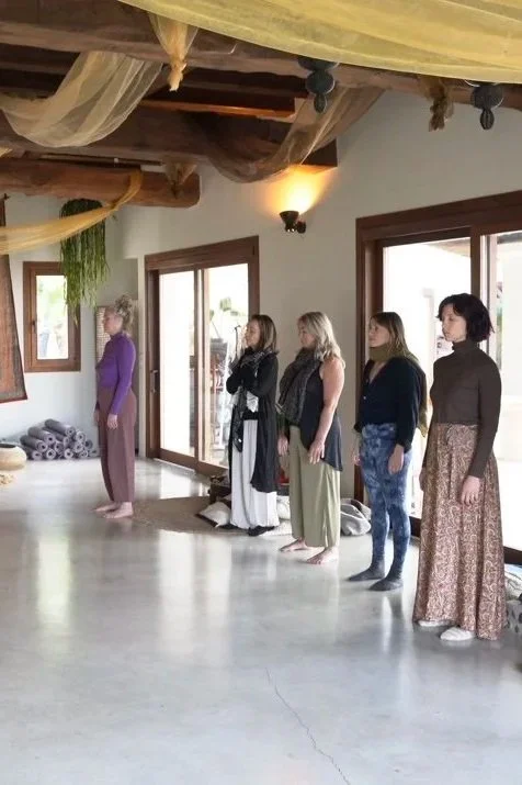 Six women standing in a line inside a room with wooden ceiling beams and large windows, participating in a yoga or meditation class.